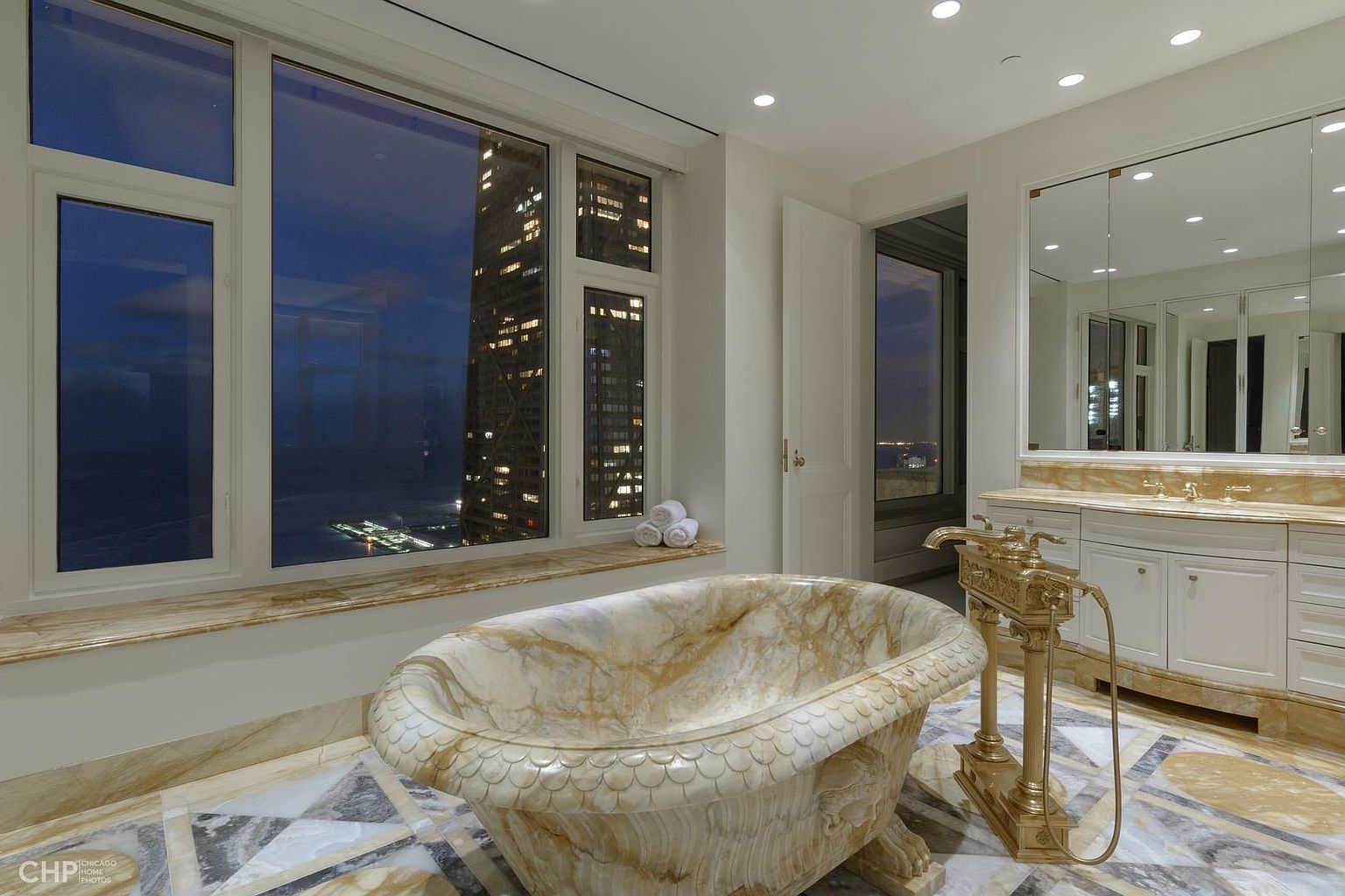 This is a luxurious primary bathroom featuring a freestanding marble bathtub with ornate gold fixtures. Large windows offer a stunning city view at night, and the room is finished with marble flooring and a vanity with ample storage and a large mirror. The overall impression is one of opulence and sophistication.