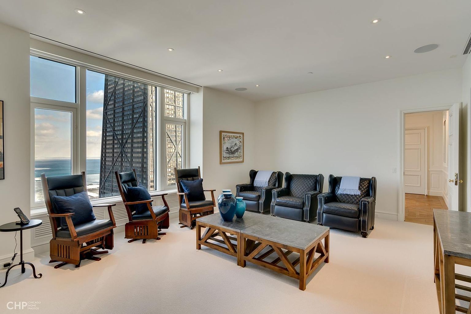 This is an interior shot of a living room featuring four leather armchairs arranged around a wooden coffee table with a stone top. The room has a neutral color palette with white walls and light carpeting. A large window offers a view of a skyscraper and the lake, while a doorway leads to another room with hardwood floors. The perspective is from the center of the room, capturing the entire seating arrangement and the view outside.