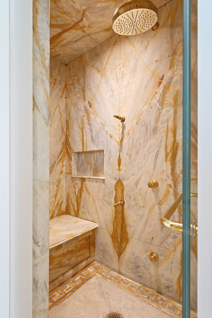 This is a luxurious primary bathroom shower featuring marble walls and a built-in bench. The shower is equipped with gold fixtures, including a rainfall showerhead and multiple body sprays. The floor has a decorative border, adding to the high-end feel of the space.