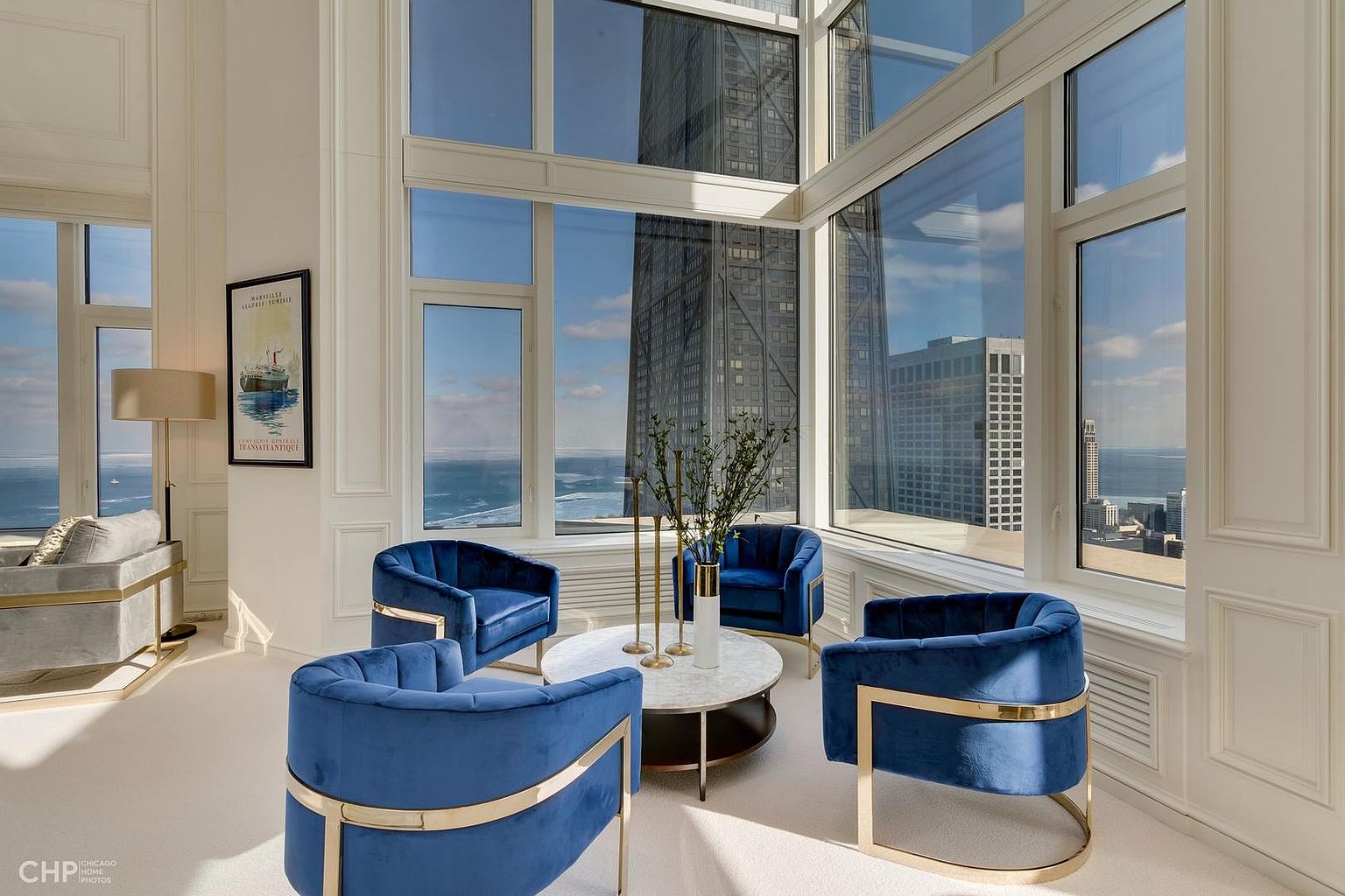 This is a luxurious living room featuring four blue velvet chairs with gold accents arranged around a marble-topped coffee table. Large windows offer stunning city views, including a prominent skyscraper. The room is bright and airy, with white walls and elegant architectural details, creating a sophisticated and inviting atmosphere.