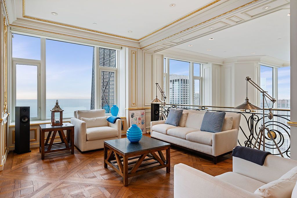 This is a luxurious living room featuring elegant architectural details such as ornate molding and large windows offering city views. The room is furnished with comfortable seating, including a sofa and armchairs, arranged around a dark wood coffee table. The space is well-lit and exudes a sophisticated and inviting atmosphere.