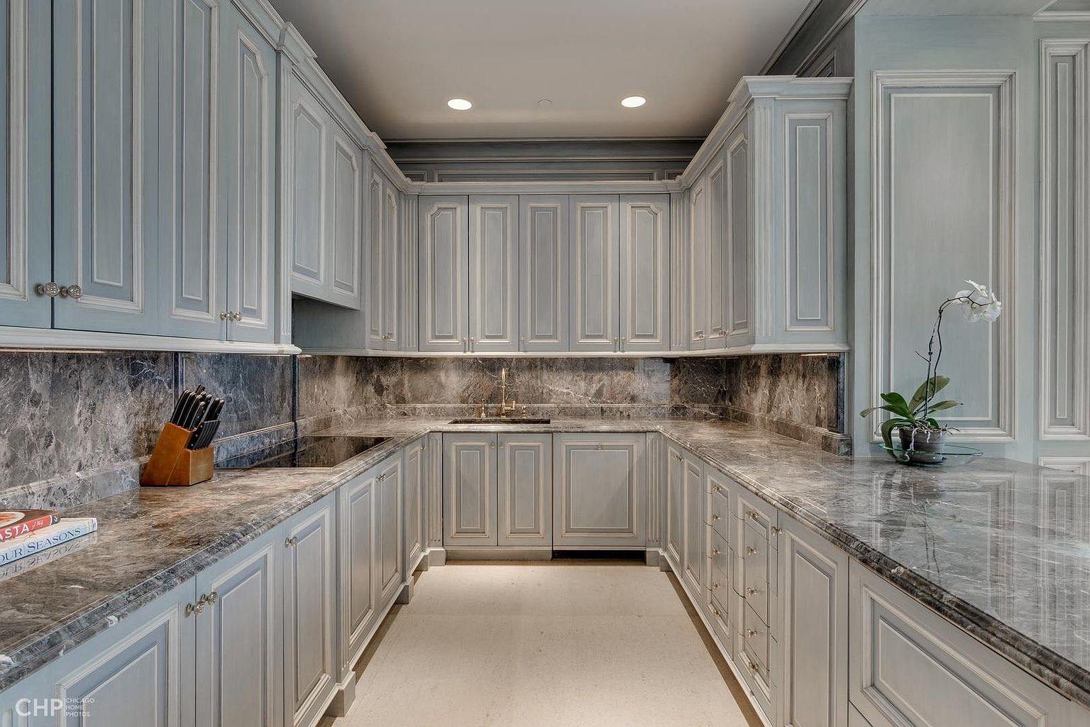 This is an interior shot of a well-appointed kitchen featuring light blue cabinetry with detailed trim and dark marble countertops and backsplash. The kitchen is arranged in a U-shape, with ample storage space provided by the numerous cabinets. The overall impression is one of elegance and sophistication, with a classic design aesthetic.