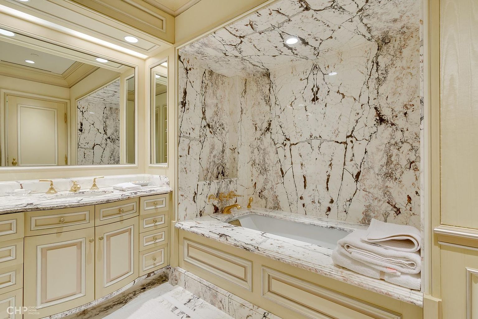 This is a luxurious primary bathroom featuring a marble-clad bathtub and walls, creating a spa-like atmosphere. The vanity area includes a large mirror and elegant cabinetry with gold fixtures. The overall design exudes sophistication and high-end quality, perfect for a discerning buyer.