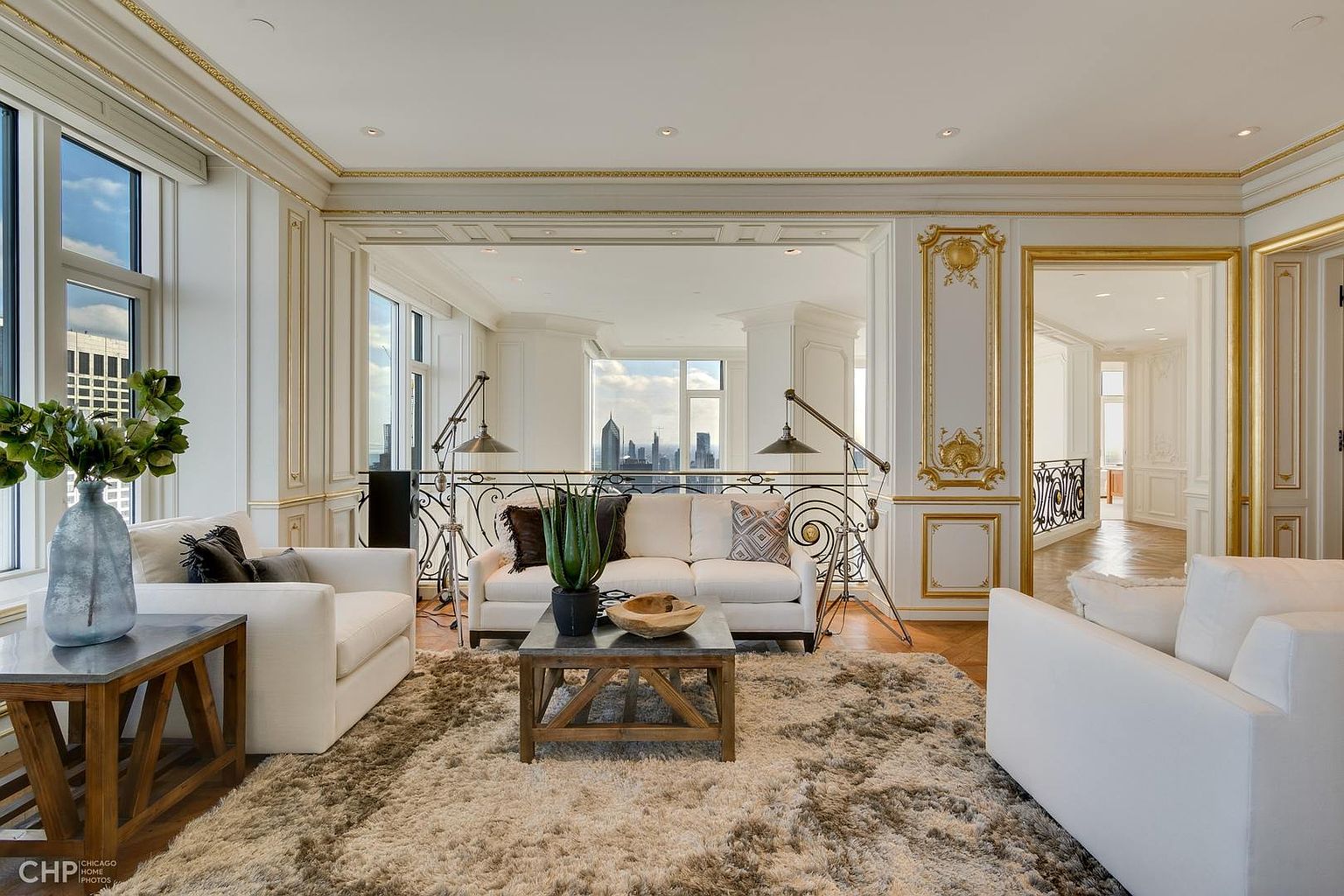 This is an interior shot of a luxurious living room featuring white sofas, a rustic wooden coffee table, and a plush shag rug. The room is adorned with ornate gold trim and large windows offering a city view. The perspective is wide, capturing the elegance and spaciousness of the room.