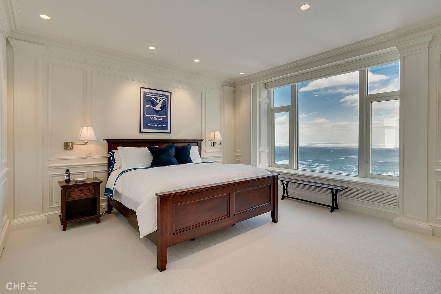 This is a primary bedroom featuring a dark wood bed with white linens and blue pillows, a matching nightstand, and a large window with a view of the ocean. The walls are paneled in white, and the room is well-lit with recessed lighting and lamps. The overall impression is luxurious and serene.