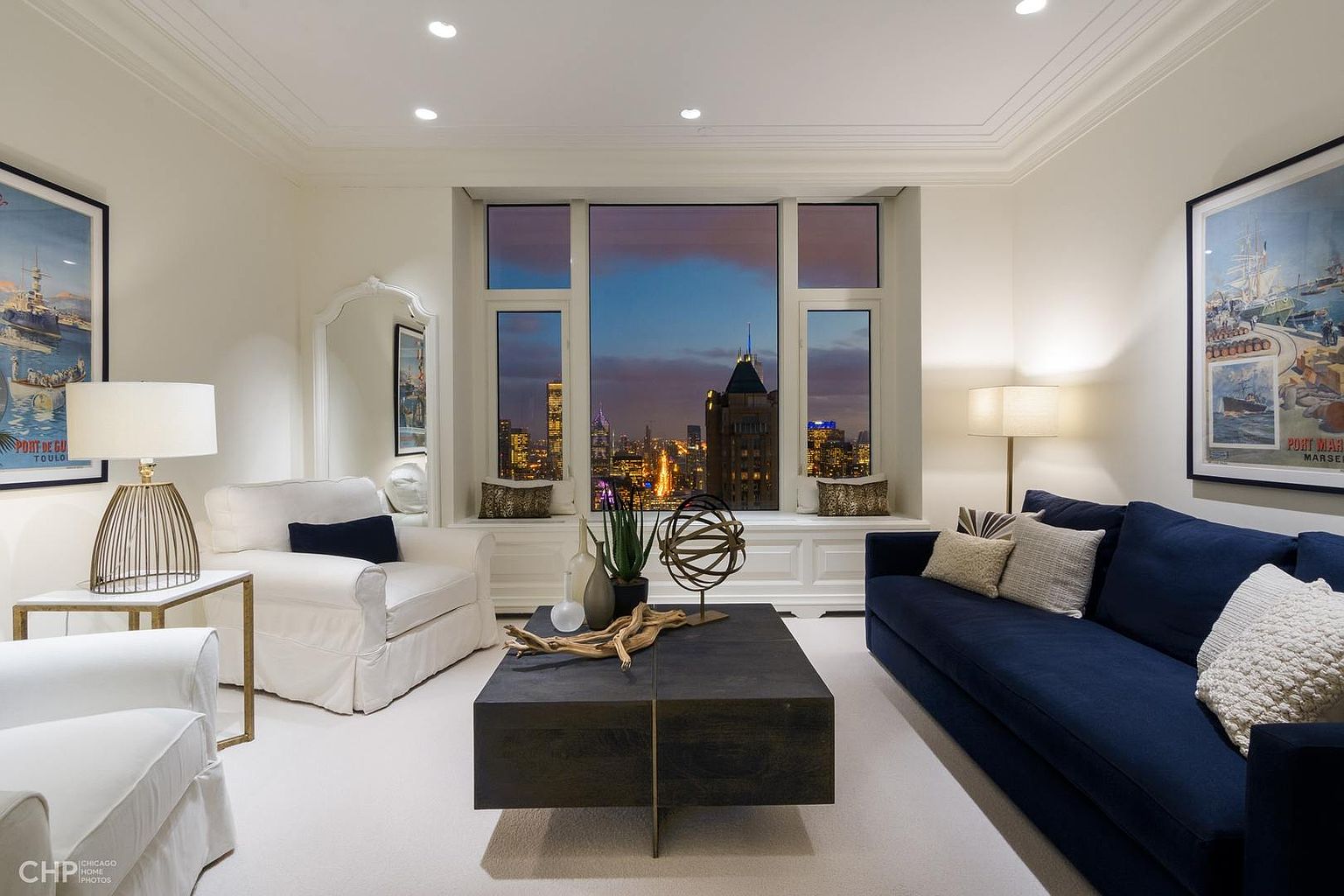 This is an interior shot of a living room featuring a large window with a city view at dusk. The room is decorated in neutral tones with a dark blue sofa, white armchairs, and a dark wood coffee table. Artwork adorns the walls, and the overall impression is one of sophisticated urban living.