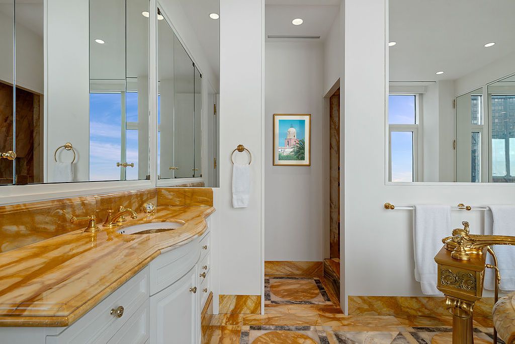 This is a luxurious primary bathroom featuring a marble countertop with gold fixtures and white cabinetry. A large mirror reflects natural light, and the floor is also marble, creating a cohesive and elegant design. The bathroom includes a separate area with a framed picture, adding a touch of sophistication.