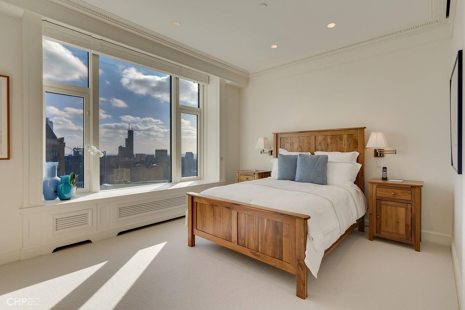 This is a bright and airy primary bedroom featuring a wooden bed frame with white linens and blue pillows. Large windows offer a stunning city view, enhancing the room's appeal. The room is carpeted and has wooden nightstands on either side of the bed.