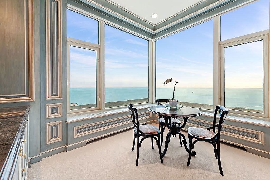 This sunroom features large windows offering panoramic views of the ocean. A small round table with chairs creates an intimate dining or relaxation space. The room is decorated with elegant, light-colored wall paneling and trim, enhancing the bright and airy atmosphere.