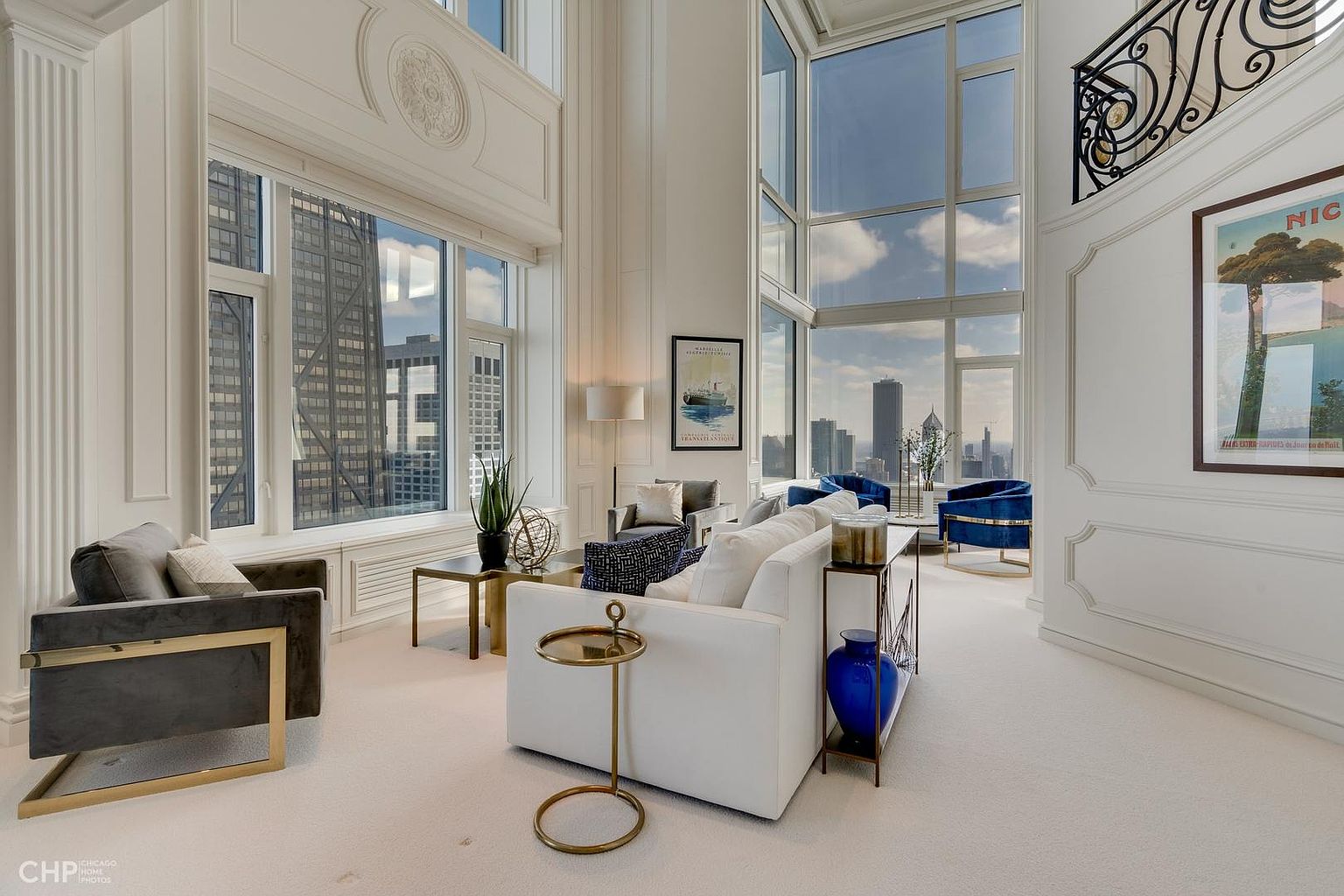 This is a luxurious living room featuring high ceilings and expansive windows offering city views. The room is furnished with modern sofas, chairs, and elegant side tables, creating a sophisticated and comfortable atmosphere. The color palette is primarily white and gray, accented with touches of gold and blue, enhancing the room's upscale appeal.