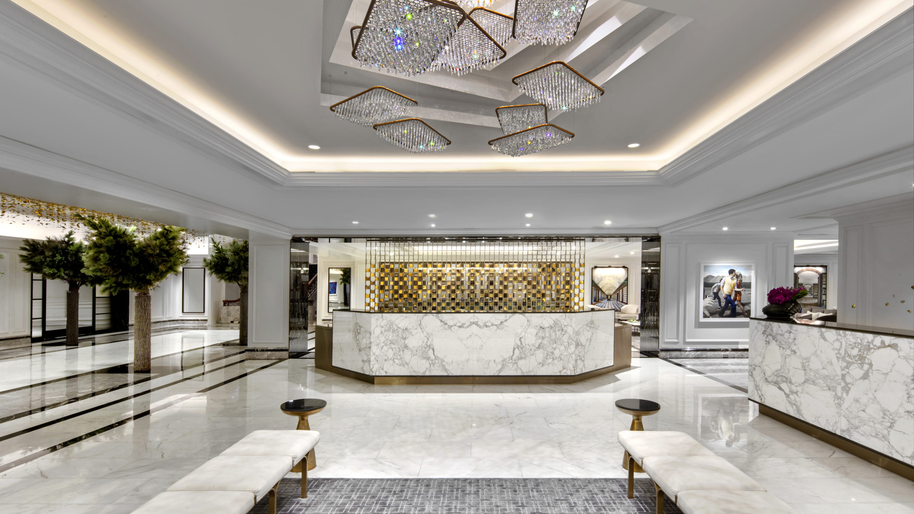 This is an interior shot of a luxurious hotel lobby. The lobby features a marble floor with black accents, elegant chandeliers, and white marble reception desks. Lush greenery adds a touch of nature to the opulent space.
