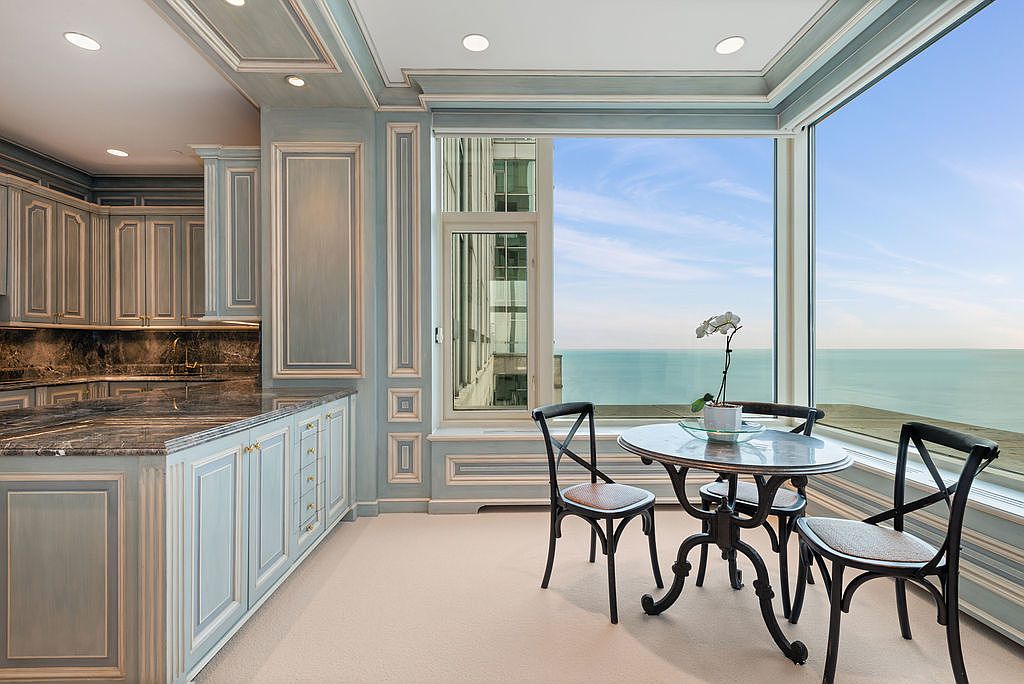 This interior shot showcases a well-appointed kitchen with custom blue cabinetry, granite countertops, and gold hardware. A breakfast nook with a round table and chairs is situated by a large window offering panoramic views of the water. The overall design exudes elegance and sophistication, perfect for a luxury real estate listing.