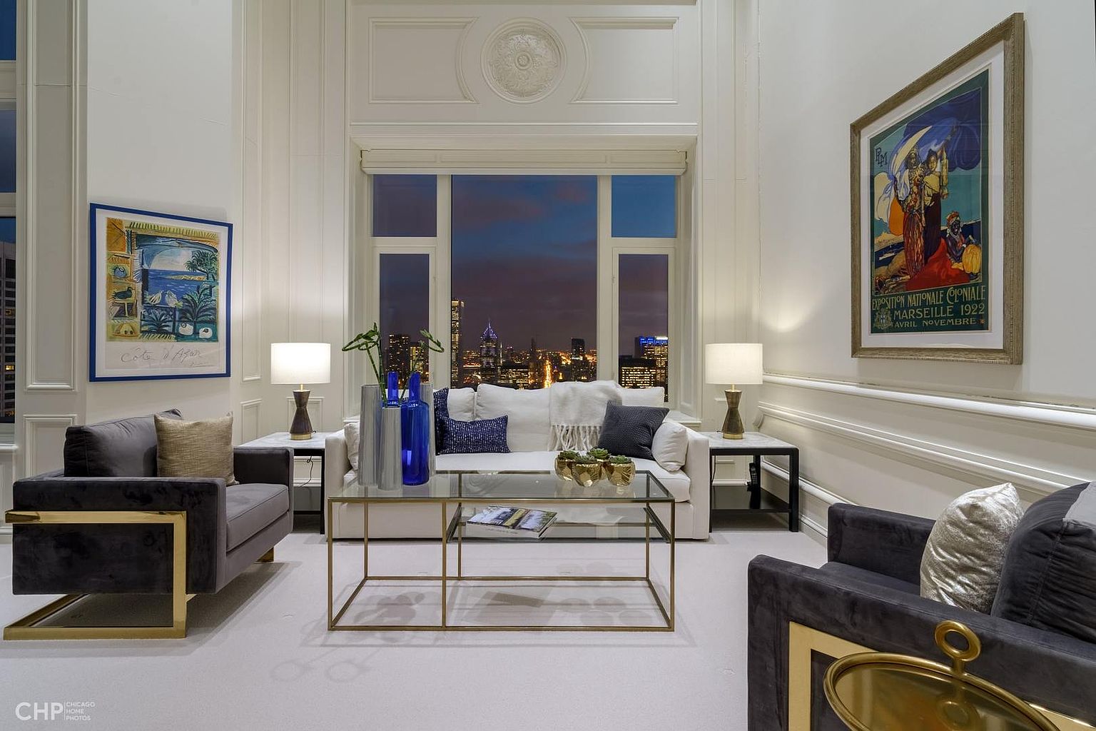 This is an elegant living room featuring a neutral color palette with pops of blue and gold accents. The room is well-lit with natural light from a large window showcasing a city skyline at dusk, complemented by table lamps. The furniture includes plush armchairs and a white sofa, arranged around a glass-topped coffee table, creating a sophisticated and inviting space.