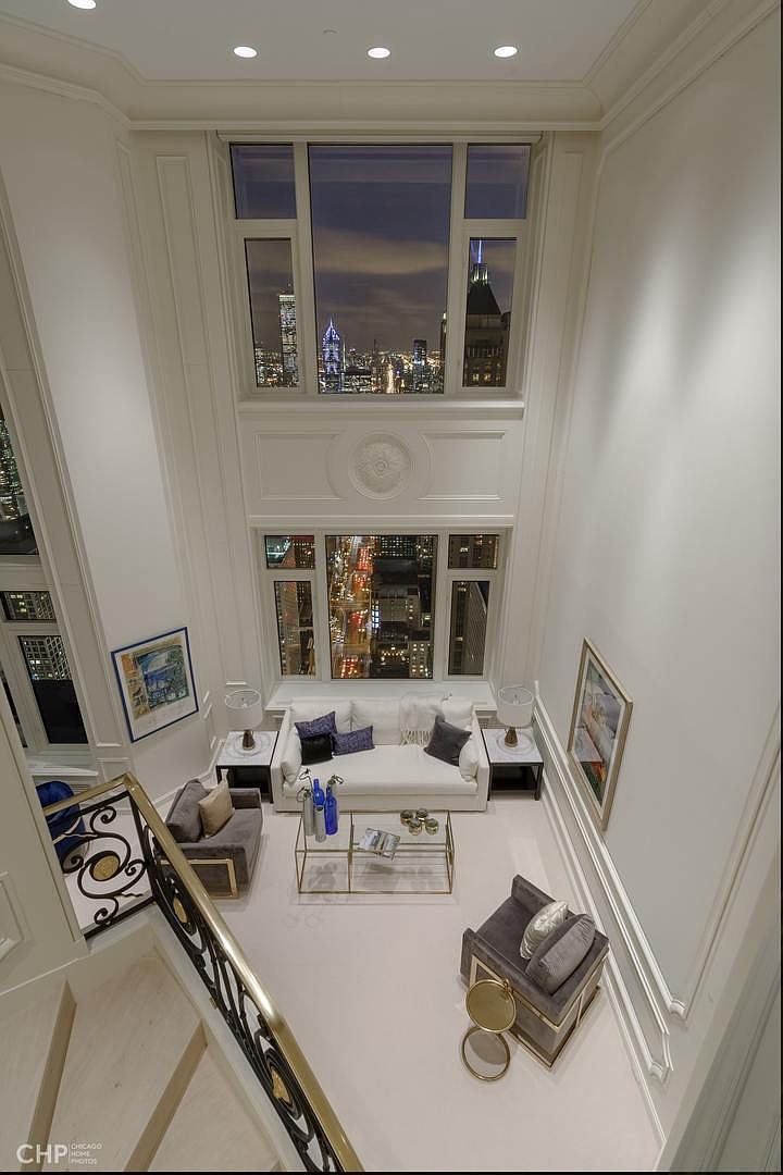 This is a high-angle shot of a luxurious living room featuring a soaring ceiling and expansive windows offering a stunning city view. The room is elegantly furnished with a white sofa, gray armchairs, and a glass coffee table, all set on a light-colored carpet. The decor is sophisticated and minimalist, emphasizing the height and light of the space.