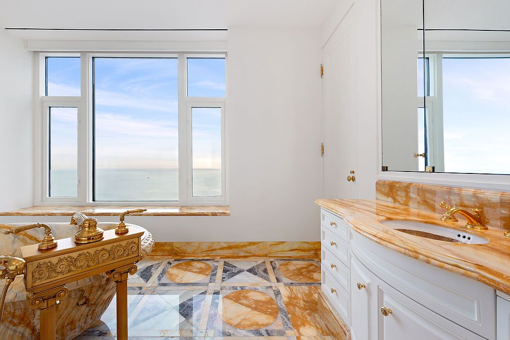 This is a luxurious primary bathroom featuring a freestanding bathtub with ornate gold fixtures positioned in front of a large window offering a scenic view. The vanity boasts a marble countertop and white cabinetry, complemented by a large mirror. The floor is tiled with a unique pattern, adding to the room's opulent feel.