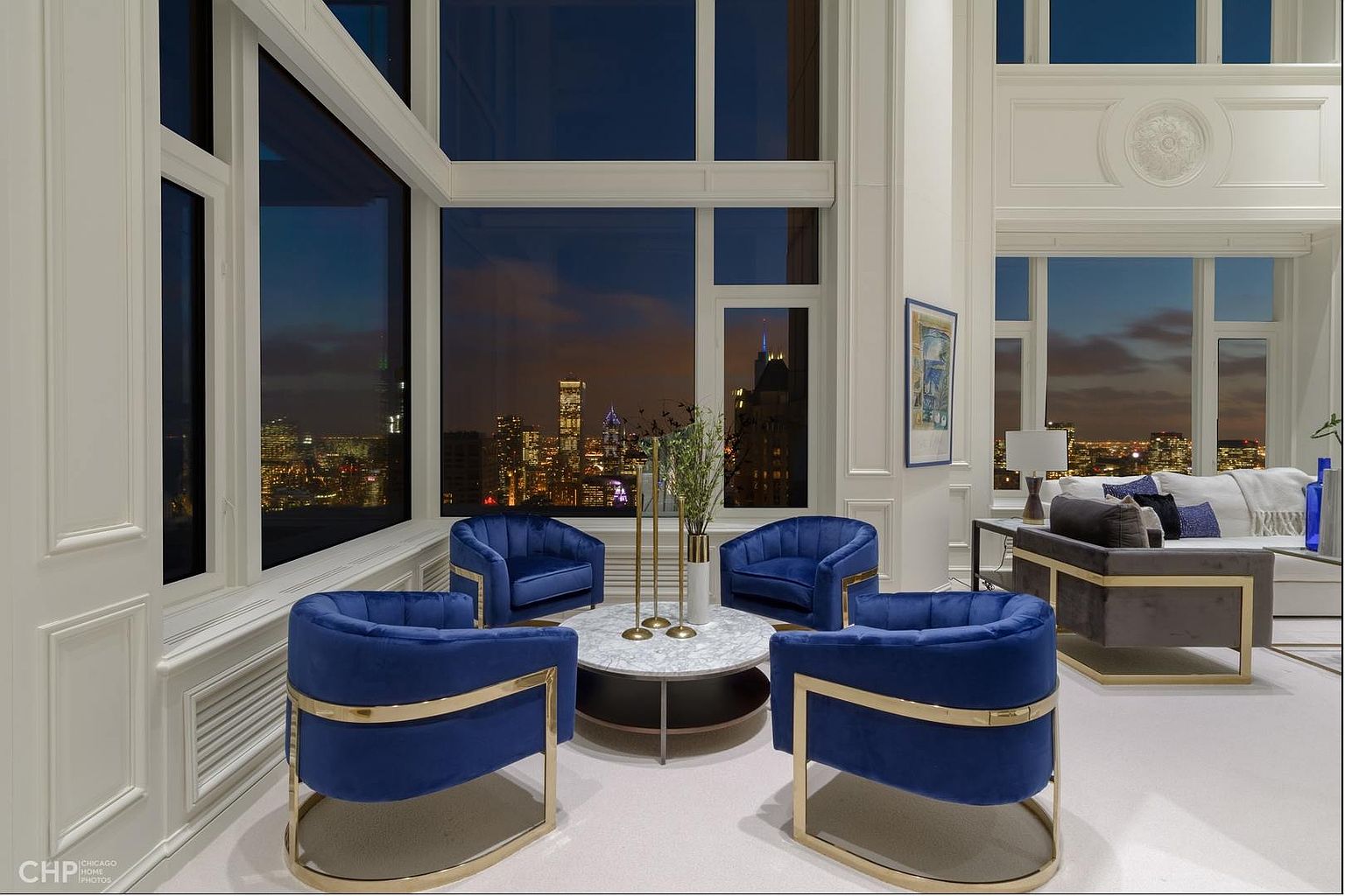This is a luxurious living room featuring four blue velvet chairs with gold accents arranged around a marble-topped coffee table. Large windows offer a stunning city view at night, and the room is decorated with elegant white wall paneling and trim. The overall impression is one of sophistication and modern elegance.
