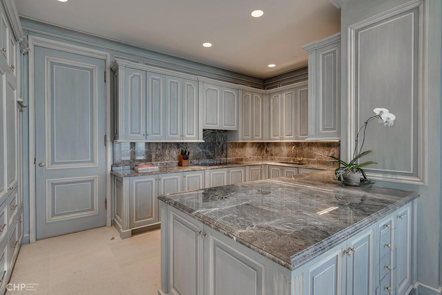 This is a well-lit kitchen featuring custom light blue cabinetry with detailed trim. A large island with a gray marble countertop dominates the center of the room, complemented by matching countertops and backsplash along the walls. The kitchen exudes a luxurious and classic style, perfect for a high-end property.