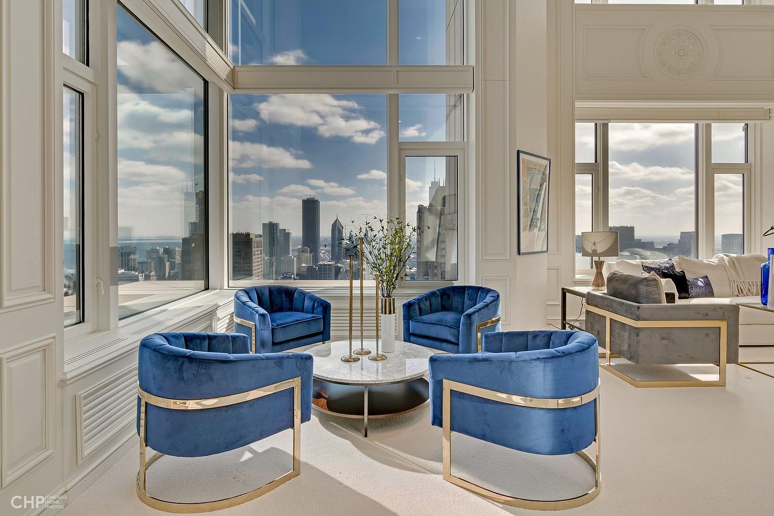 This is an interior shot of a luxurious living room featuring four blue velvet chairs with gold accents arranged around a round marble coffee table. Large windows offer a stunning city view, enhancing the room's bright and airy atmosphere. The space is elegantly decorated with modern furniture and neutral tones, creating a sophisticated and inviting ambiance.