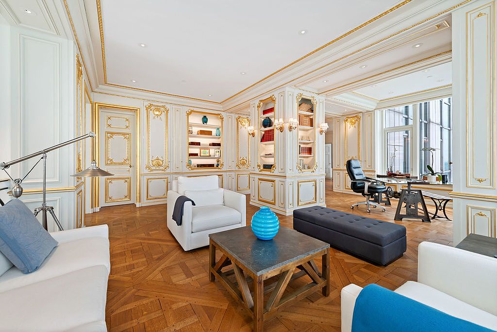 This is an interior shot of a luxurious office or study, featuring ornate white and gold paneling, built-in bookshelves, and hardwood floors in a herringbone pattern. The room is furnished with white armchairs, a dark ottoman, and a rustic coffee table, creating a blend of classic elegance and modern comfort. A large window provides natural light, highlighting the room's sophisticated design.
