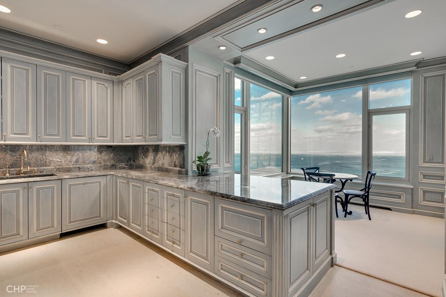 This is a high-end kitchen with custom gray cabinetry, marble countertops, and stainless steel appliances. The kitchen features a large island and a breakfast nook with floor-to-ceiling windows offering stunning ocean views. The overall impression is luxurious and sophisticated.