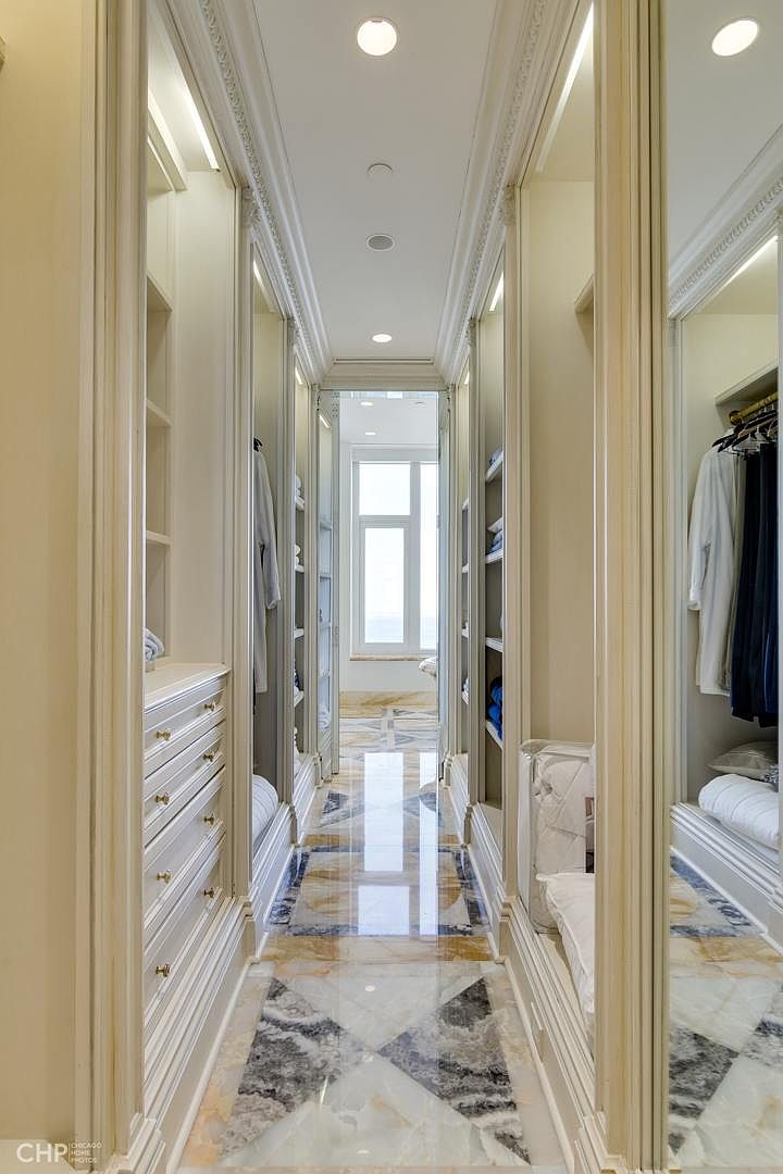 This is a luxurious walk-in closet featuring custom cabinetry, drawers, and shelving. The closet has a long, narrow layout with mirrored doors and a marble floor with a geometric pattern. The space is well-lit with recessed lighting and accent lights, creating an elegant and organized atmosphere.