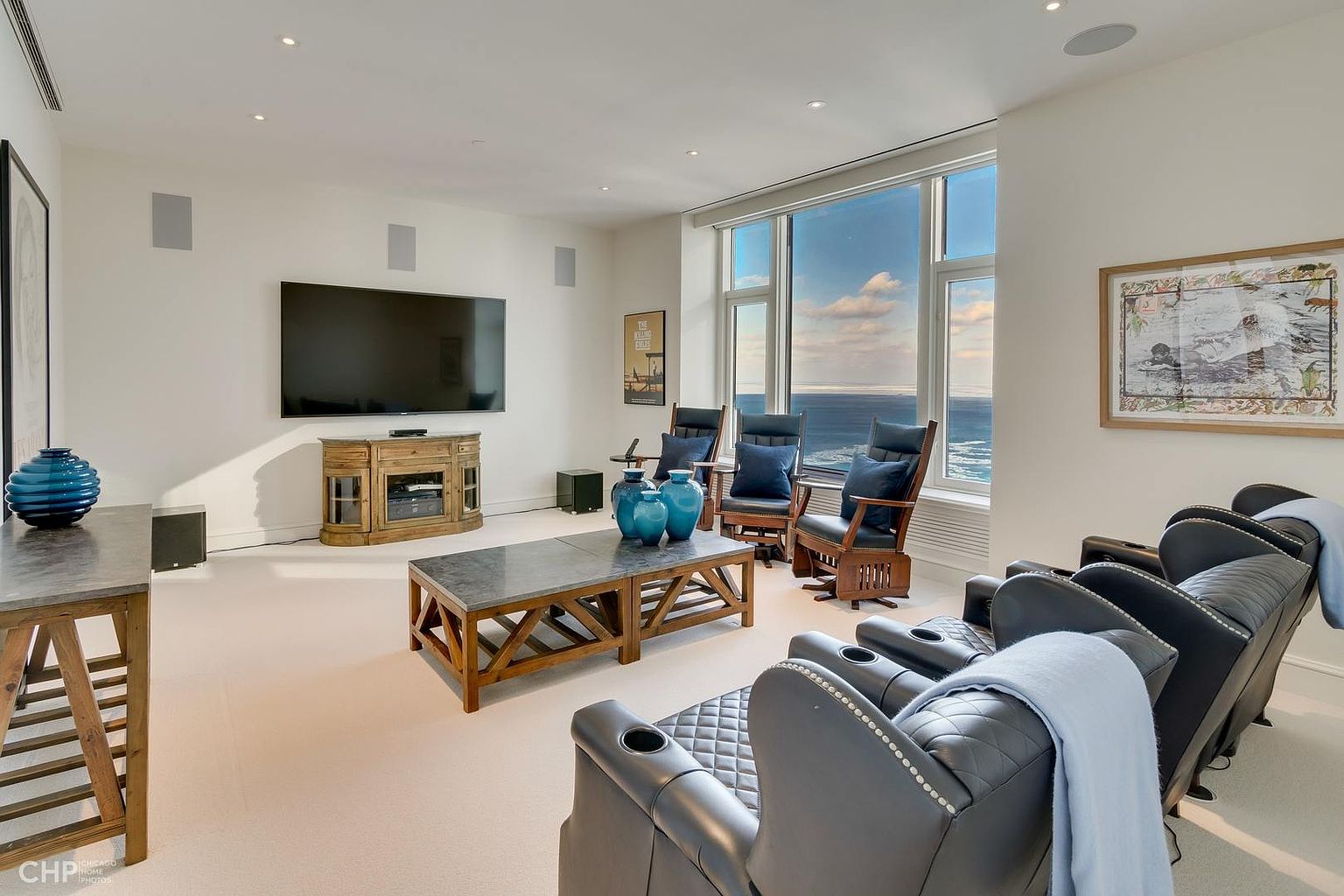 This is a well-lit living room featuring a large flat-screen TV mounted on the wall above a wooden media console. The room is furnished with comfortable-looking leather reclining chairs, a wooden coffee table with a stone top, and additional seating near a large window with a view of the ocean. The overall aesthetic is modern and luxurious, creating a relaxing and inviting space.