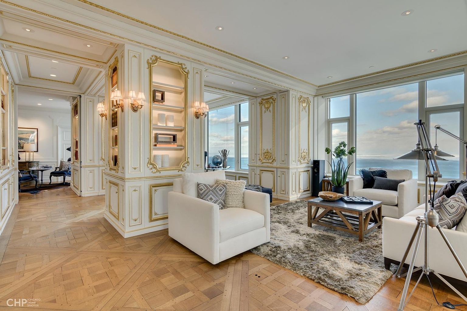 This is a luxurious living room featuring ornate white and gold paneling, large windows with ocean views, and elegant furnishings. The room is bathed in natural light, highlighting the hardwood floors and plush area rug. The overall impression is one of sophistication and opulence, perfect for high-end real estate.
