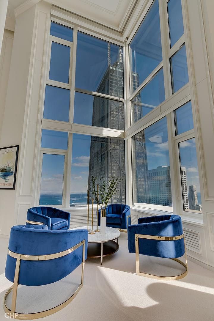 This is an interior shot of a luxurious living room featuring four blue velvet chairs with gold accents arranged around a round coffee table. The room boasts floor-to-ceiling windows offering a stunning view of a skyscraper and the city skyline. The overall impression is one of modern elegance and sophistication.