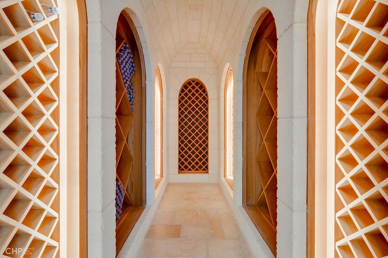 This is an interior shot of a wine cellar featuring arched alcoves with wooden wine racks. The walls and ceiling are constructed of light-colored stone, creating a rustic yet elegant ambiance. The floor is tiled with large, rectangular stones, and the lighting is soft and warm, highlighting the architectural details and the wine storage.