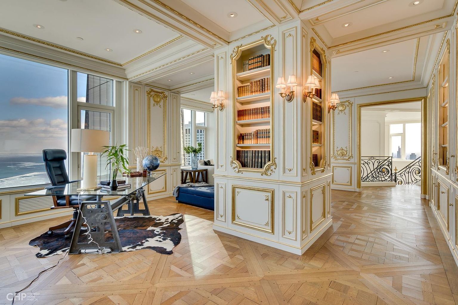 This is a luxurious home office featuring custom white and gold built-in bookshelves, a glass-top desk with a unique metal base, and a cowhide rug. The room is bathed in natural light from a large window offering a city view, and the herringbone wood flooring adds to the sophisticated ambiance. The space exudes elegance and functionality, perfect for a discerning professional.