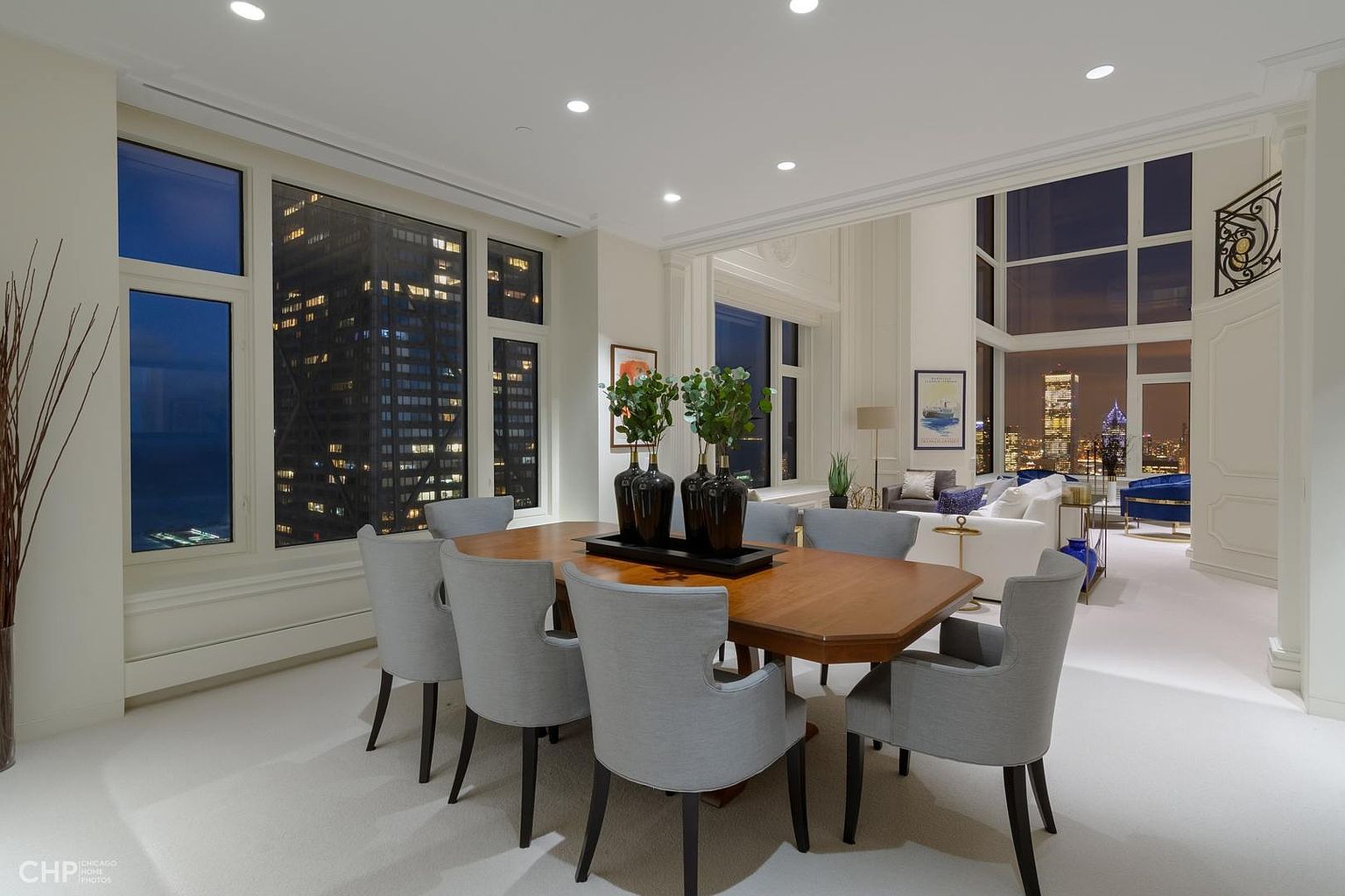 This is a luxurious dining room featuring a large wooden table surrounded by six gray upholstered chairs. The room is bright with natural light from large windows offering a stunning city view at night. The space flows seamlessly into a living area, creating an open and elegant atmosphere.