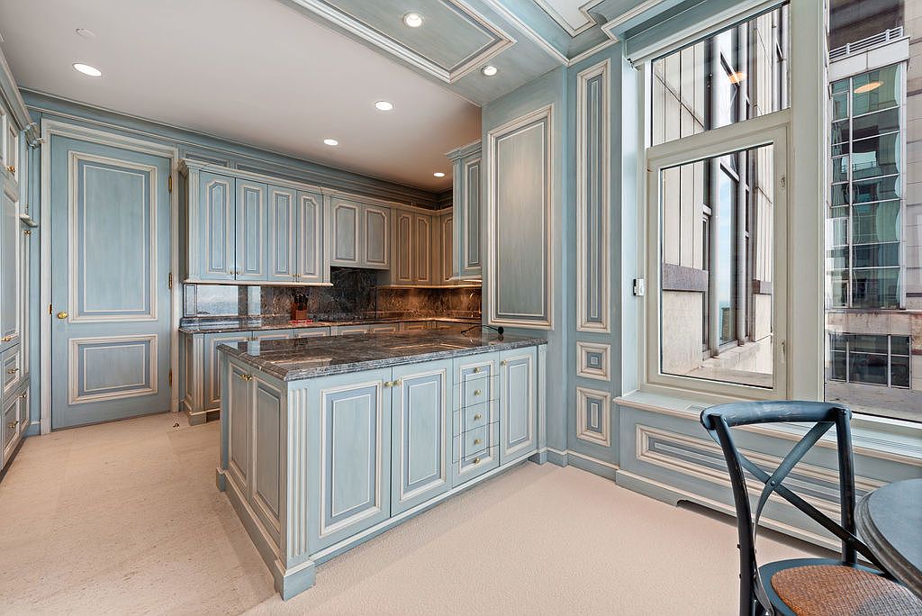 This is a well-lit kitchen featuring custom blue cabinetry with white trim, granite countertops, and a central island. The kitchen has a traditional style with ornate detailing and a large window offering a city view. The space appears luxurious and well-maintained.