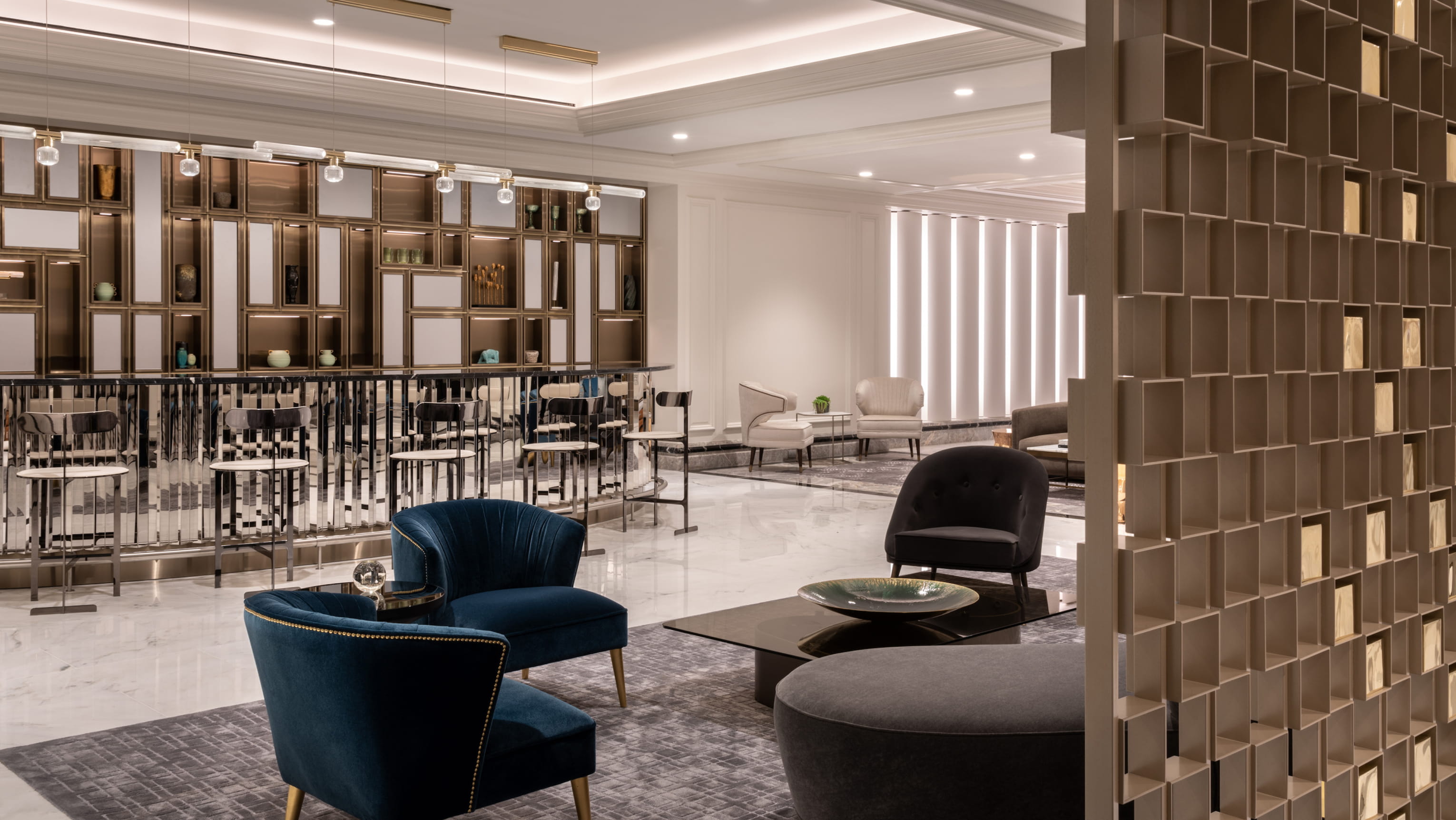This interior showcases a stylish living room area with plush blue armchairs, a modern coffee table, and a gray ottoman. The space features a sophisticated bar area in the background and is accented with a light, neutral color palette. The design conveys an atmosphere of luxury and comfort, suitable for a high-end residential property or hotel lobby.
