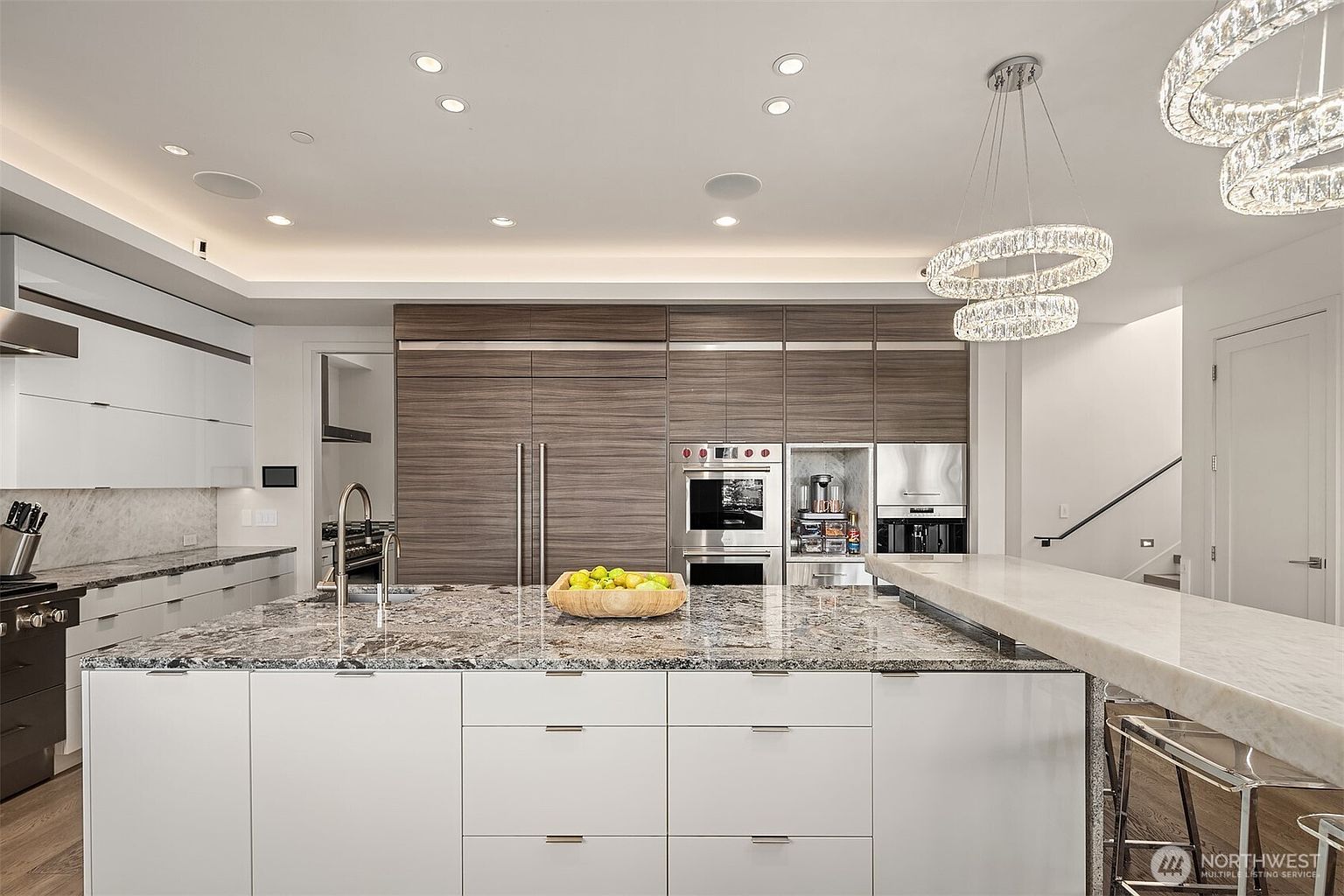 This modern kitchen features a large central island with a stone countertop and a wooden bowl of fruit, serving as the focal point of the room. The space is equipped with sleek white lower cabinetry and a wall of dark wood-toned cabinetry that houses built-in stainless steel appliances. Two ornate, circular crystal chandeliers hang above the island, while recessed lighting and cove lighting provide a bright, sophisticated atmosphere.