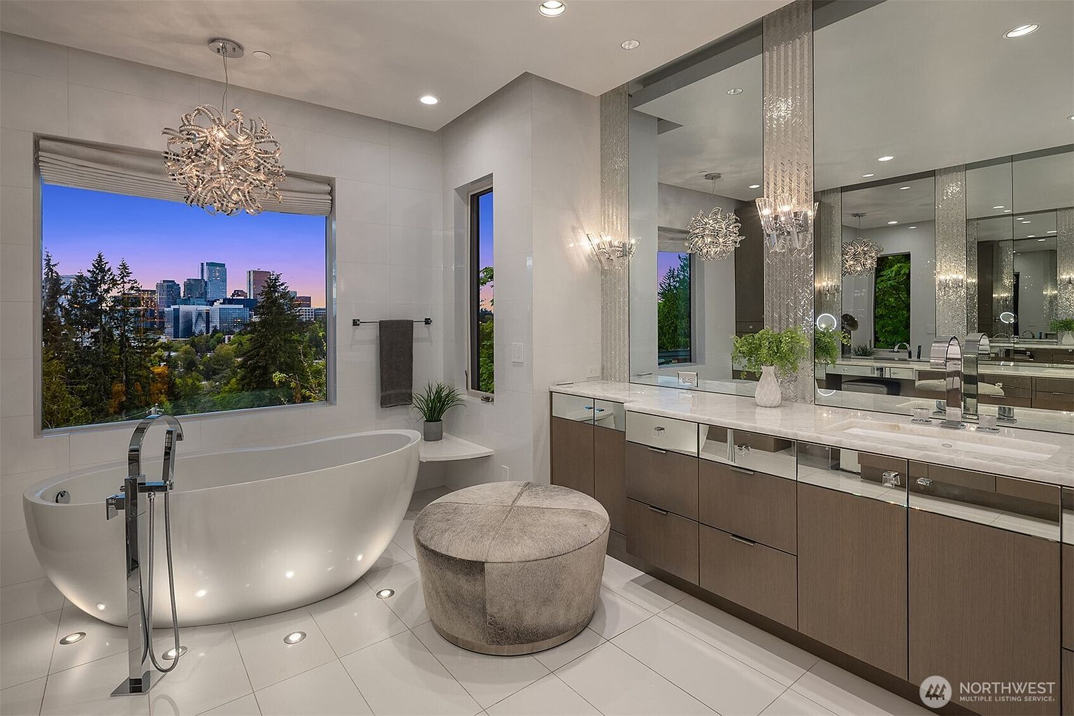 This luxurious primary bathroom features a sleek, freestanding soaking tub positioned beneath a large window that offers a panoramic view of the city skyline. The space is anchored by a long vanity with a stone countertop, mirrored cabinetry, and elegant light fixtures, all set against a backdrop of polished tile flooring. A plush, circular ottoman sits in the center, enhancing the sophisticated and serene atmosphere of the room.