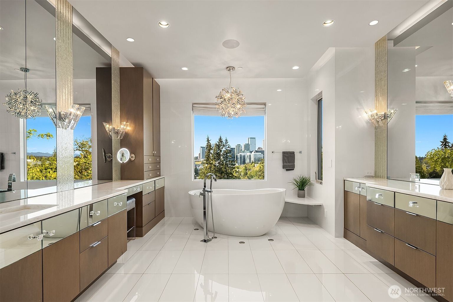 This luxurious primary bathroom features a central freestanding soaking tub positioned in front of a large window with a panoramic city skyline view. The space is symmetrically designed with dual vanities, sleek wood-toned cabinetry, and large mirrors accented by decorative crystal-style light fixtures. The room is finished with polished light-colored tile flooring and recessed ceiling lighting, creating a bright and sophisticated atmosphere.