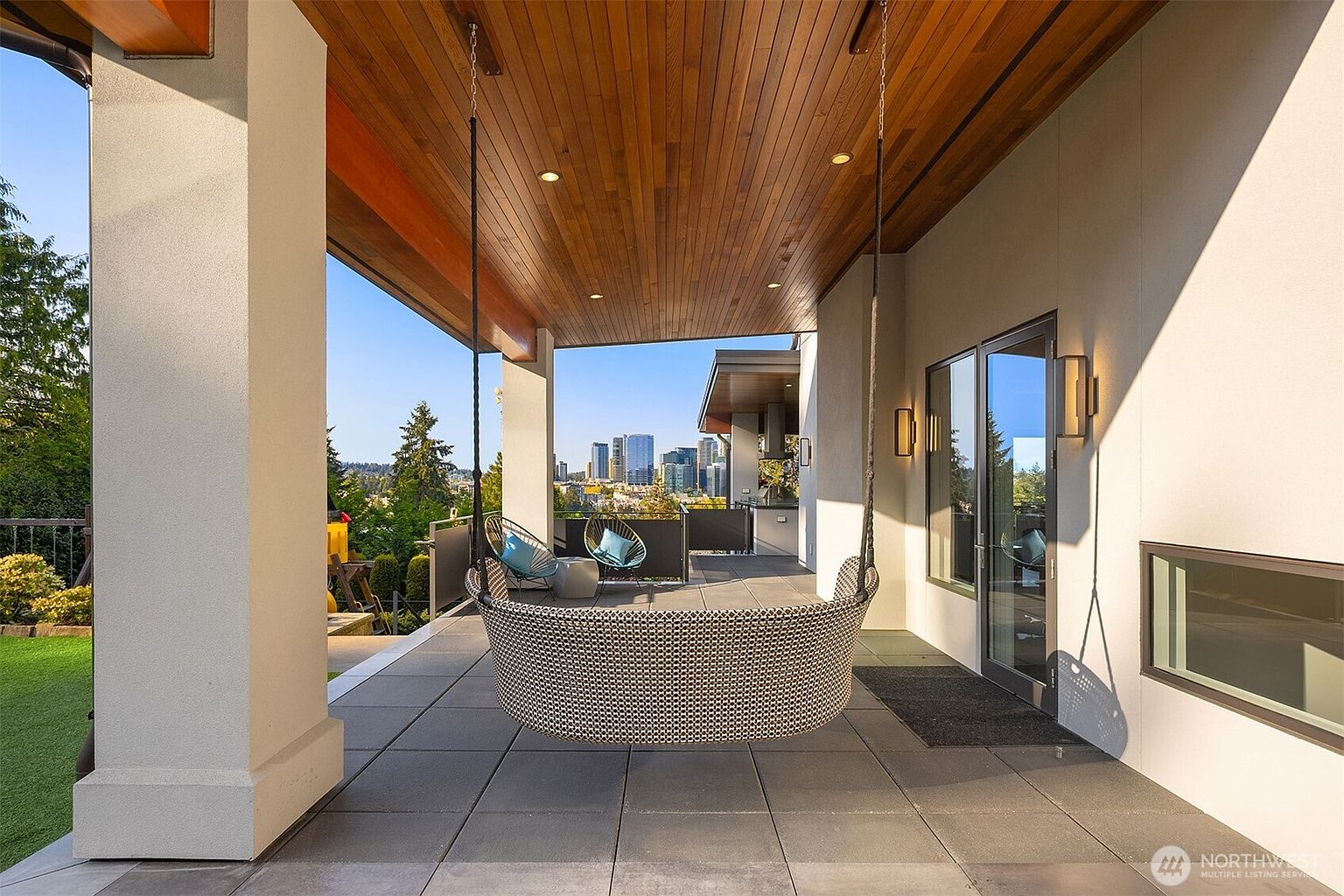 This modern covered patio features a large, suspended woven swing chair as a central focal point, set against a backdrop of a stunning city skyline view. The space is finished with dark square floor tiles, a warm wood-paneled ceiling with recessed lighting, and sleek glass railings. Two additional outdoor chairs are positioned further out on the deck, creating a sophisticated and inviting outdoor living area.