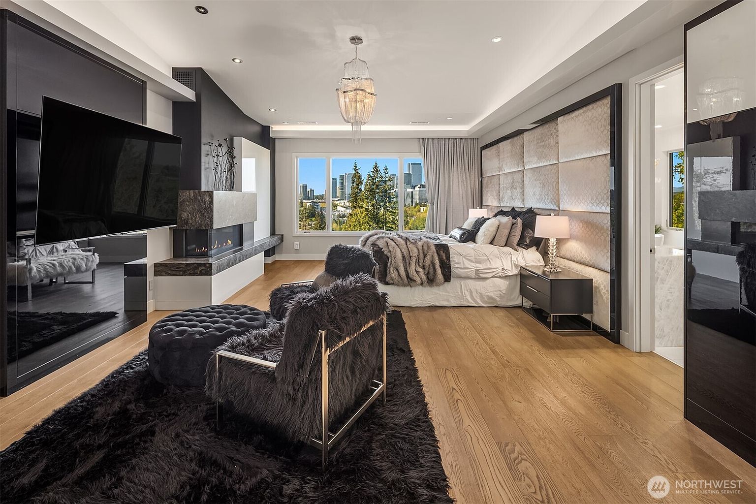 This luxurious primary bedroom features a large bed centered against a tall, upholstered headboard wall with a grid pattern. A cozy seating area with two dark, textured armchairs and a matching ottoman sits on a plush black rug, while a modern fireplace with a stone surround adds warmth to the space. Large windows provide an expansive view of the city skyline, and the room is finished with light wood flooring and a sophisticated crystal chandelier.