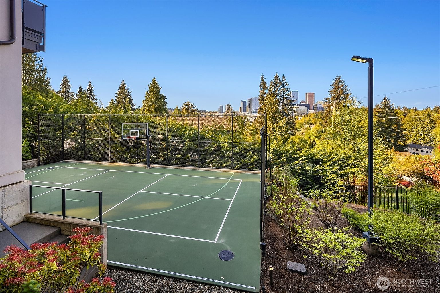 This elevated outdoor space features a private, fenced-in sports court with a basketball hoop, set against a backdrop of lush trees and a distant city skyline. The perspective is from an upper-level vantage point, looking down onto the green court surface and the surrounding landscaped garden area. The scene conveys a sense of luxury and recreation, highlighting the property's unique combination of private sports facilities and scenic urban views.