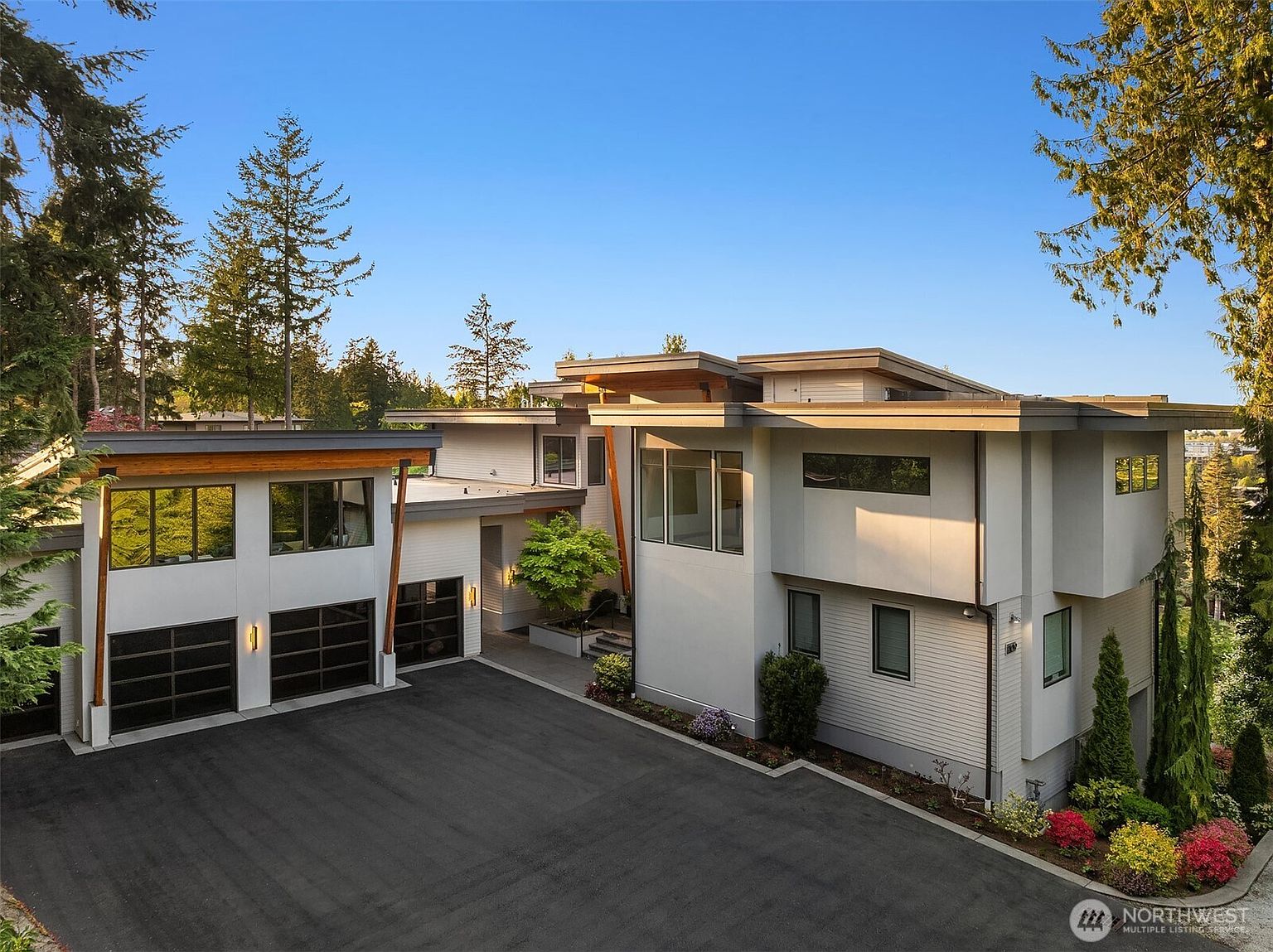 This modern, multi-level home features a striking contemporary design with clean lines, a mix of light-colored siding, and warm wood accents. The property includes a spacious paved driveway leading to a three-car garage, with a recessed entryway tucked between the garage and the main living structure. Large windows and geometric architectural forms create a sophisticated, high-end aesthetic set against a backdrop of mature evergreen trees.