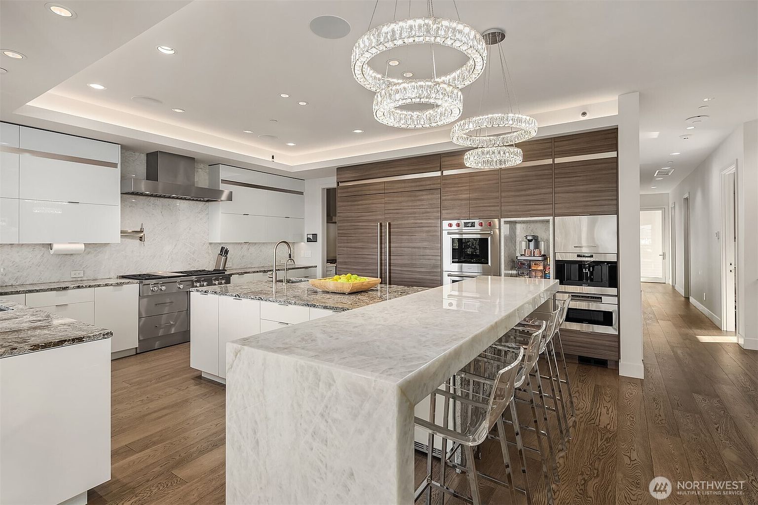 This modern, high-end kitchen features a large central island with a waterfall-edge stone countertop and bar seating. The space is defined by a contrast between sleek white cabinetry and warm-toned wood cabinetry, complemented by professional-grade stainless steel appliances and dual crystal chandeliers. The perspective captures the expansive layout, highlighting the seamless transition from the cooking area to the adjacent hallway.