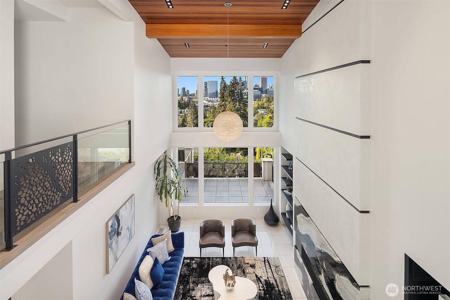 This high-ceilinged living room features a dramatic wood-paneled ceiling with recessed lighting and a large, spherical pendant light. The space is anchored by a blue velvet sofa and two leather armchairs, set against a tall, minimalist wall with horizontal black accents and a dark stone fireplace surround. Large windows provide an expansive view of the city skyline and surrounding trees, creating a bright and airy atmosphere.