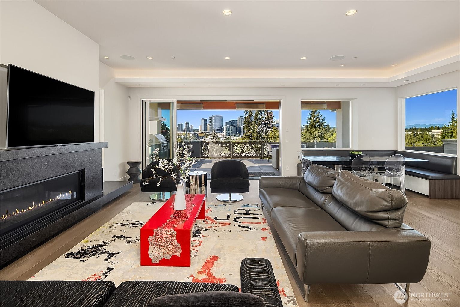 This contemporary living room features a sleek, dark-toned fireplace with a wide, linear firebox and a prominent stone-clad chimney breast. A vibrant red coffee table serves as a focal point atop a patterned area rug, complemented by a dark leather sofa and two modern armchairs. Large sliding glass doors open to a spacious balcony, framing a clear view of the city skyline and surrounding greenery.