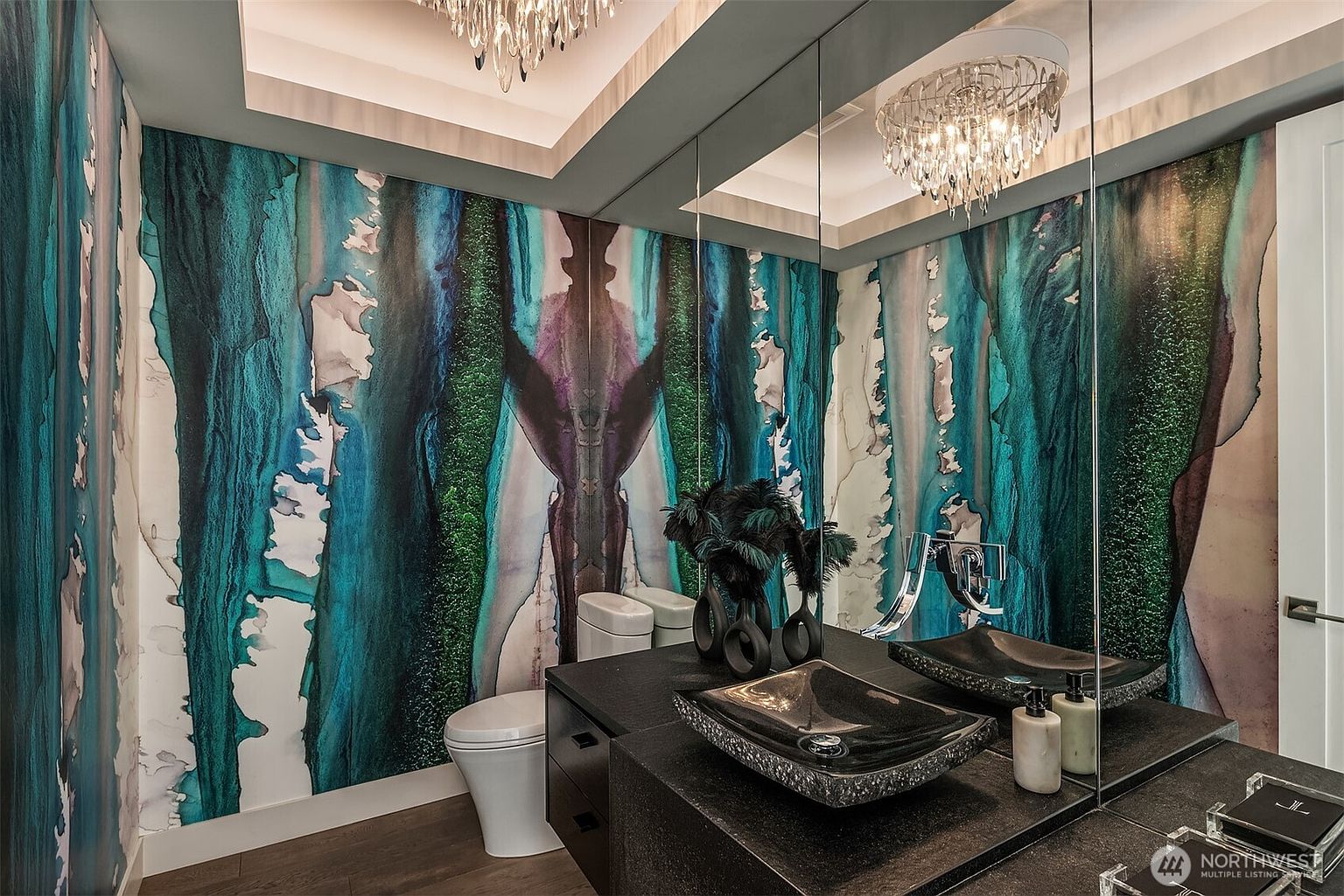 This striking powder room features bold, abstract wallpaper in deep teal and purple tones that creates a dramatic focal point. A dark, textured vanity holds a unique, dark-toned vessel sink, complemented by a large mirror that reflects the space and the intricate crystal chandelier above. The room exudes a luxurious and moody aesthetic, enhanced by the sophisticated lighting and high-contrast design elements.
