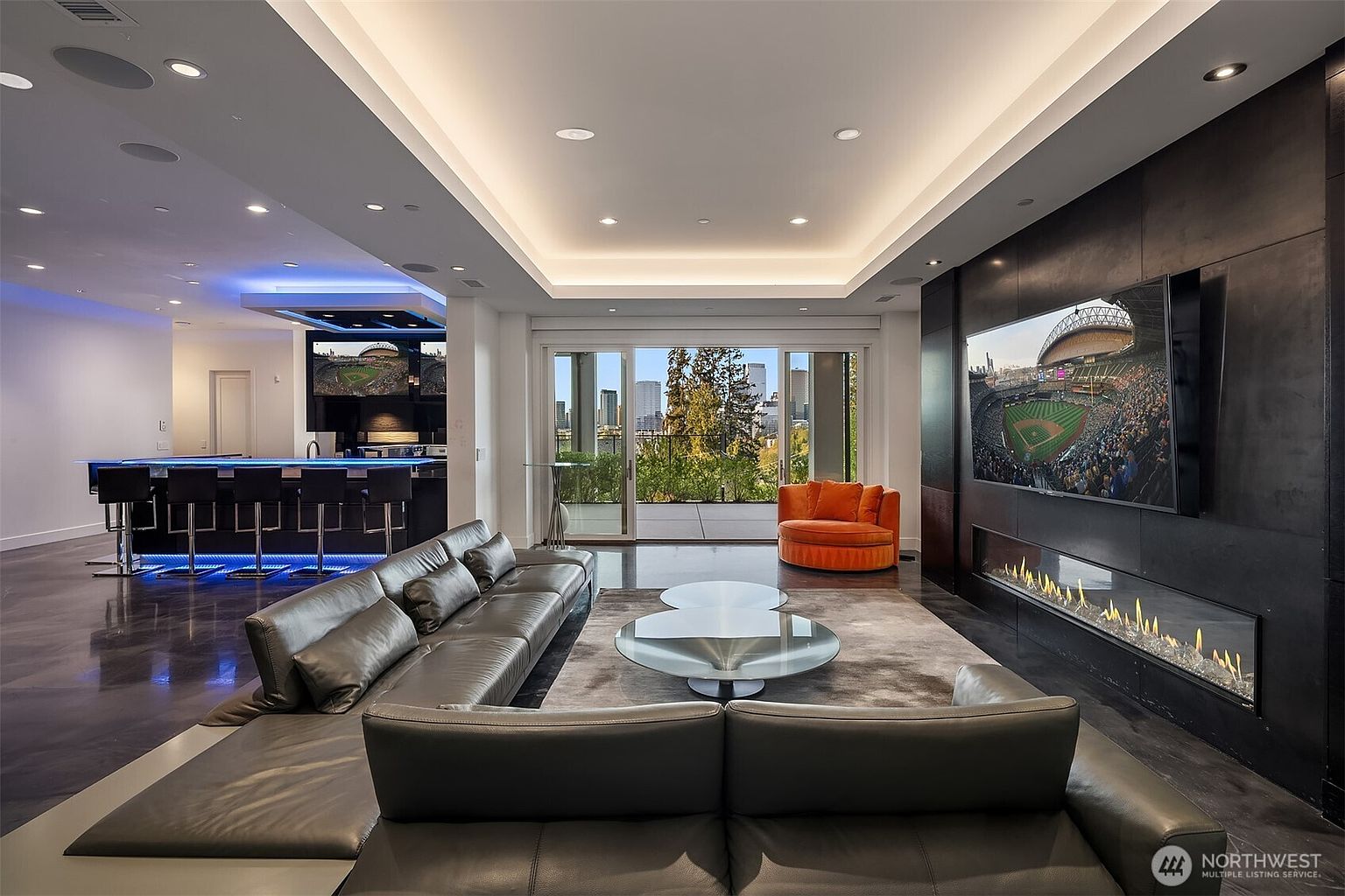 This expansive, modern living room features a sleek, dark-toned media wall with a wide, linear fireplace and a large mounted television. A spacious, dark leather sectional sofa faces the media wall, centered around a unique glass-topped coffee table on a plush area rug. To the left, a contemporary bar area with blue accent lighting and bar stools creates an ideal space for entertaining, while large sliding glass doors open to a balcony with a city skyline view.