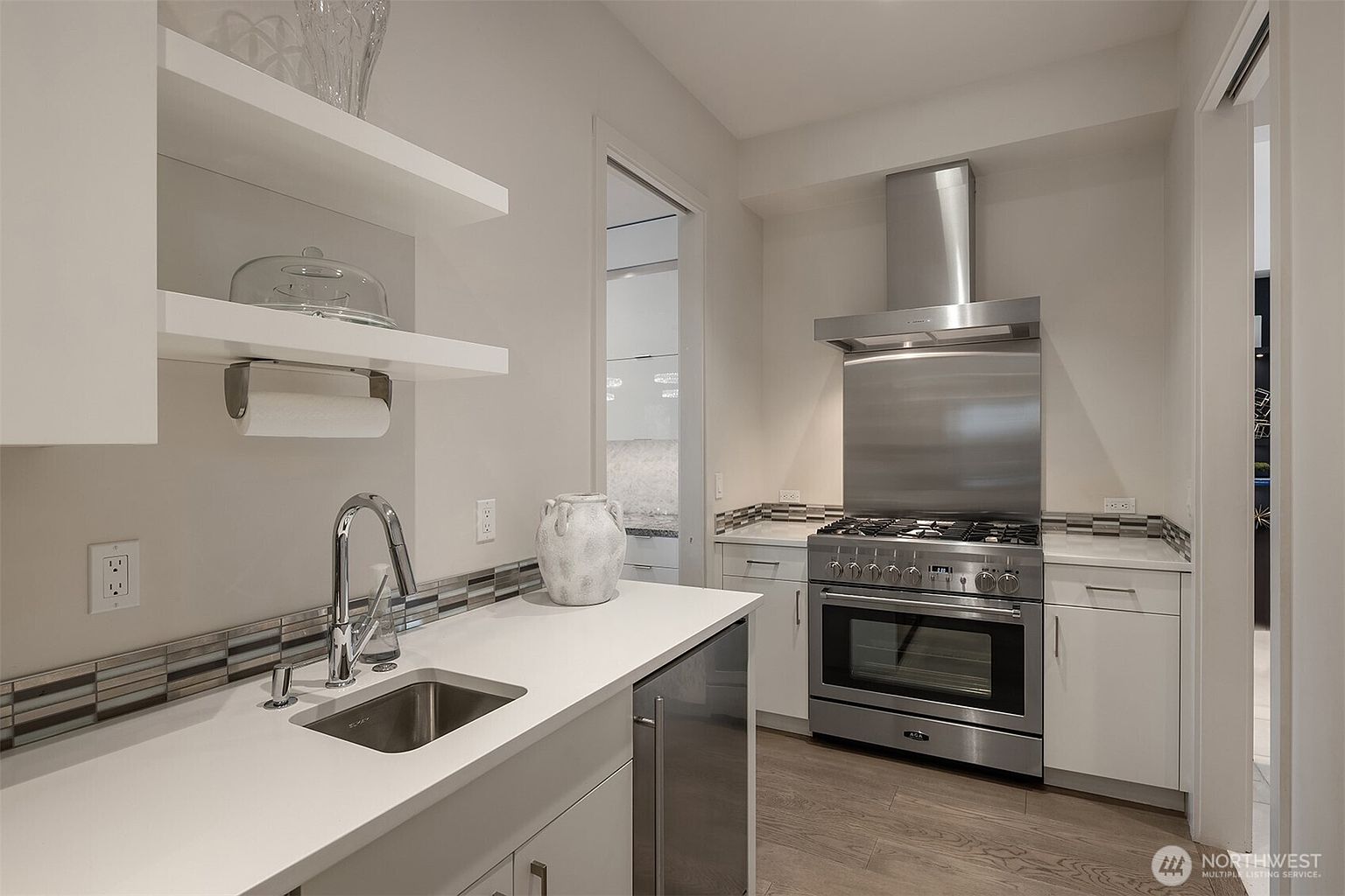 This modern kitchenette features a sleek stainless steel gas range with a matching vent hood and a compact under-counter sink area. The space is finished with light-toned cabinetry, a patterned tile backsplash, and light wood-look flooring, creating a clean and functional aesthetic. A doorway in the background provides a glimpse into an adjacent room, adding depth to the composition.