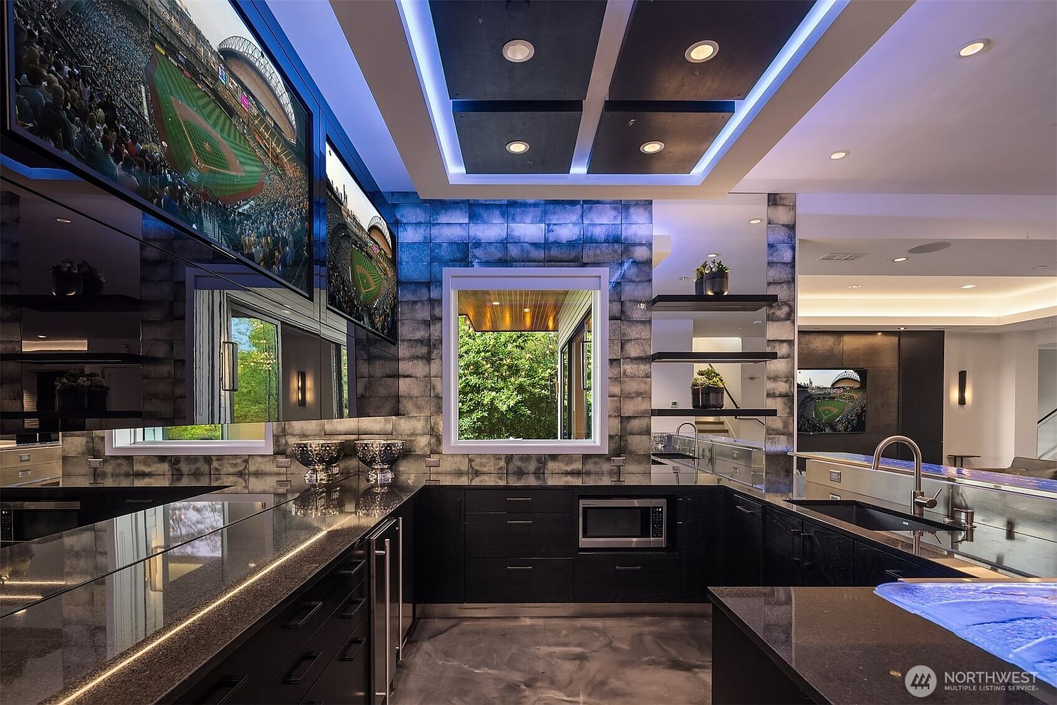 This modern, high-end kitchen features dark cabinetry, sleek stone countertops, and a unique tiled wall backsplash that adds texture to the space. The room is accented by vibrant blue LED lighting integrated into the ceiling panels and under the counter, creating a sophisticated, lounge-like atmosphere. Multiple screens displaying sports games are mounted above the counters, emphasizing the space's entertainment-focused design.