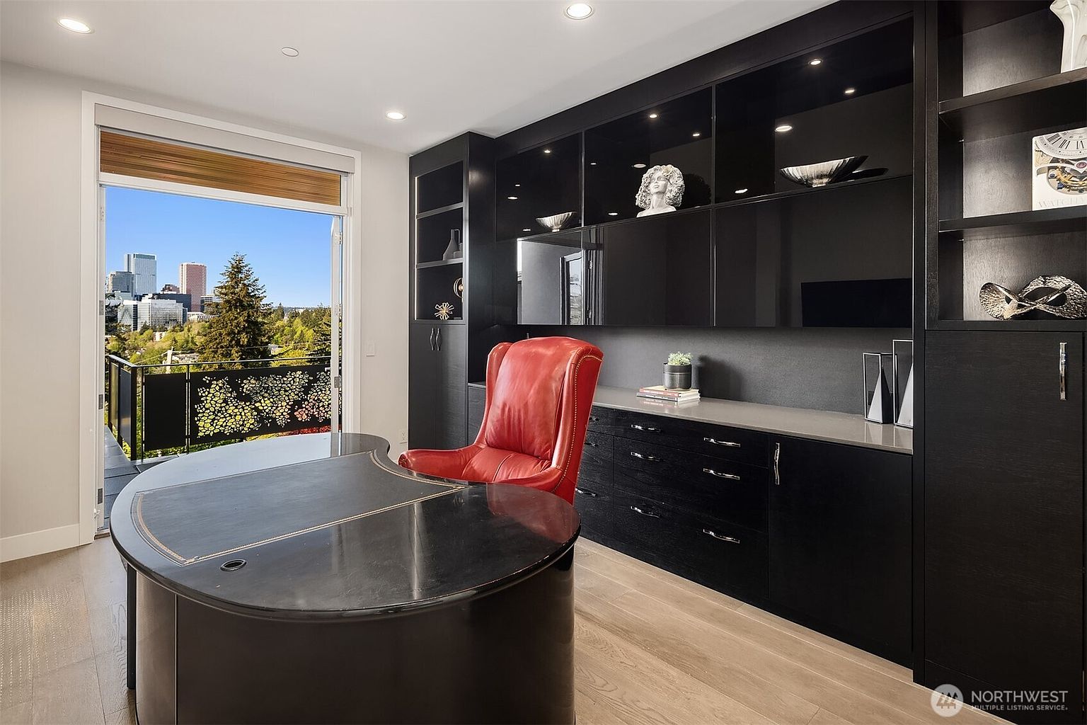 This sophisticated home office features a striking contrast between dark, high-gloss cabinetry and light wood flooring. A bold red leather chair sits behind a curved, dark-toned desk, while a large glass door opens to a balcony with an expansive city skyline view. The room is accented with decorative sculptures and recessed lighting, creating a professional yet modern atmosphere.