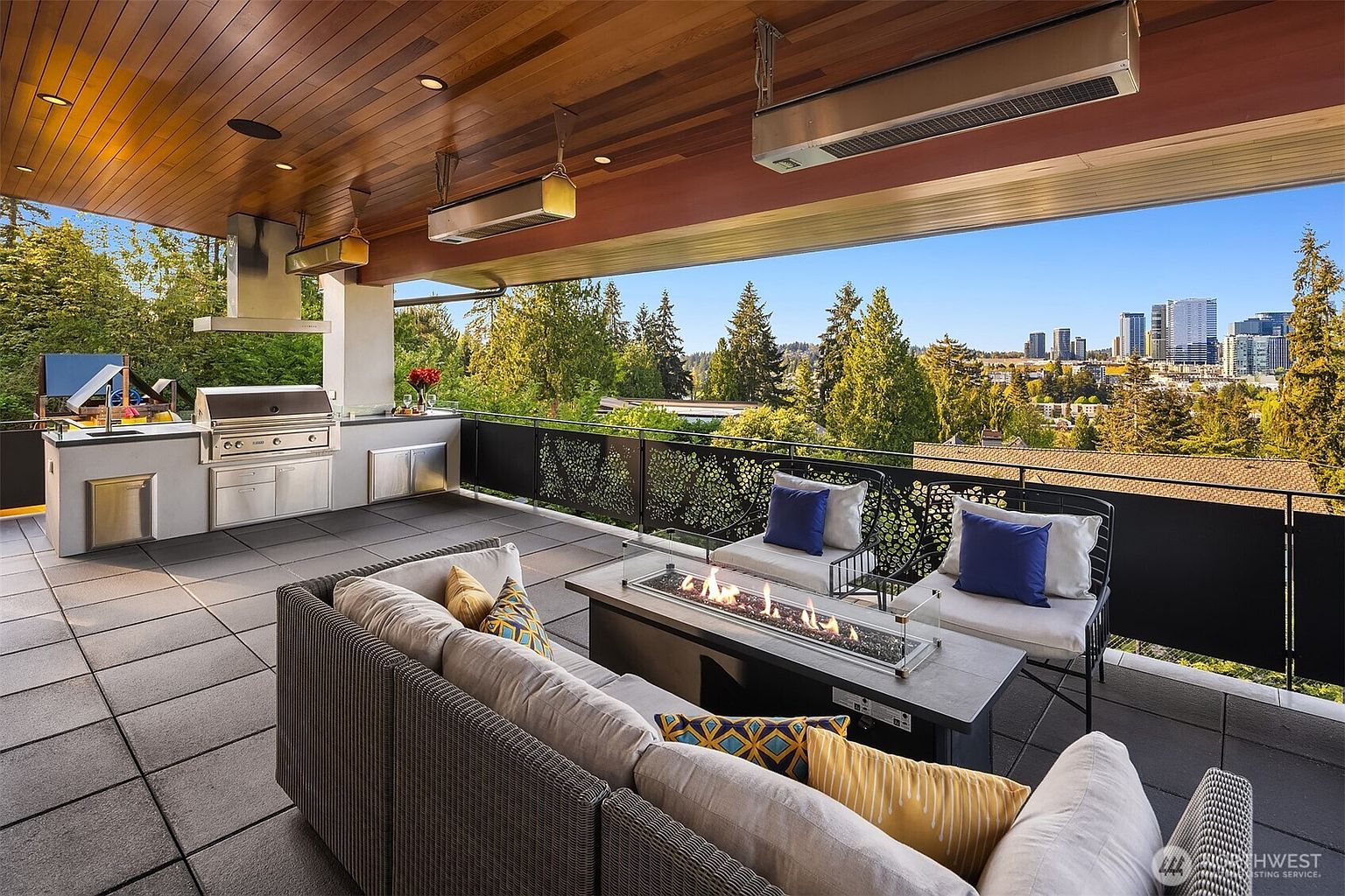 This expansive covered balcony offers a sophisticated outdoor living space complete with a built-in stainless steel kitchen and a central fire table. The area is furnished with a comfortable sectional sofa and armchairs, all set against a backdrop of lush trees and a distant city skyline. The space features a warm wood-paneled ceiling with integrated lighting and overhead heaters, creating an inviting atmosphere for entertaining.