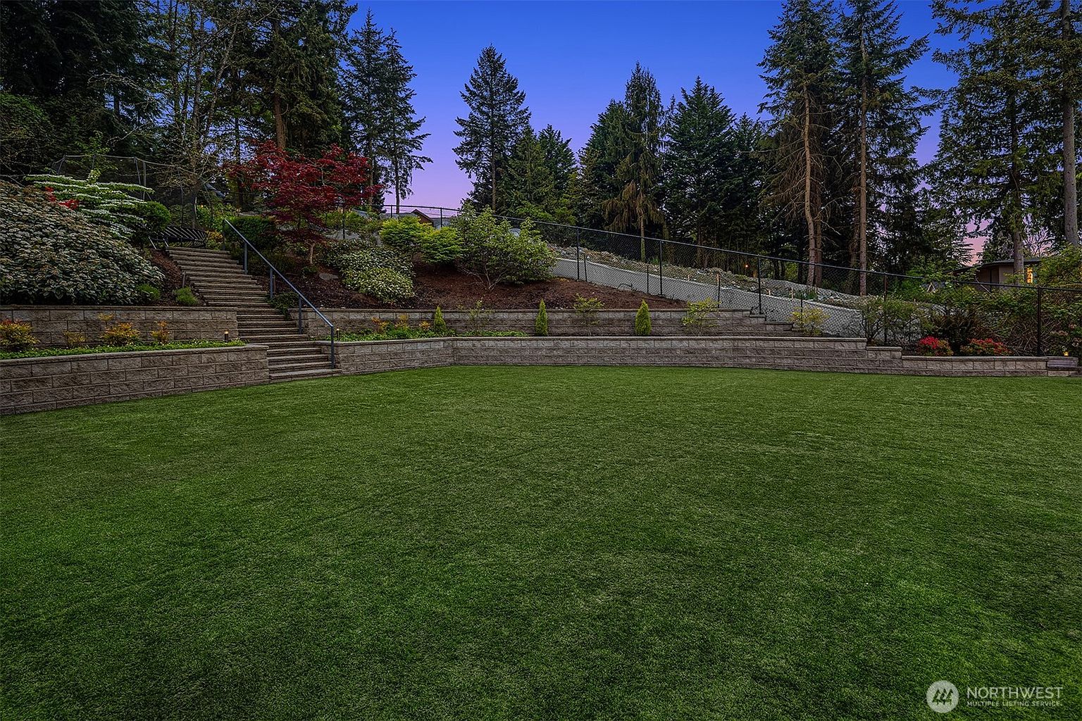 This expansive backyard features a meticulously maintained, level lawn bordered by a tiered stone retaining wall. A set of stone stairs leads up the landscaped hillside, which is densely planted with mature trees and shrubs, all enclosed by a black chain-link fence. The scene is captured at dusk, with the twilight sky providing a serene backdrop to the well-manicured outdoor space.